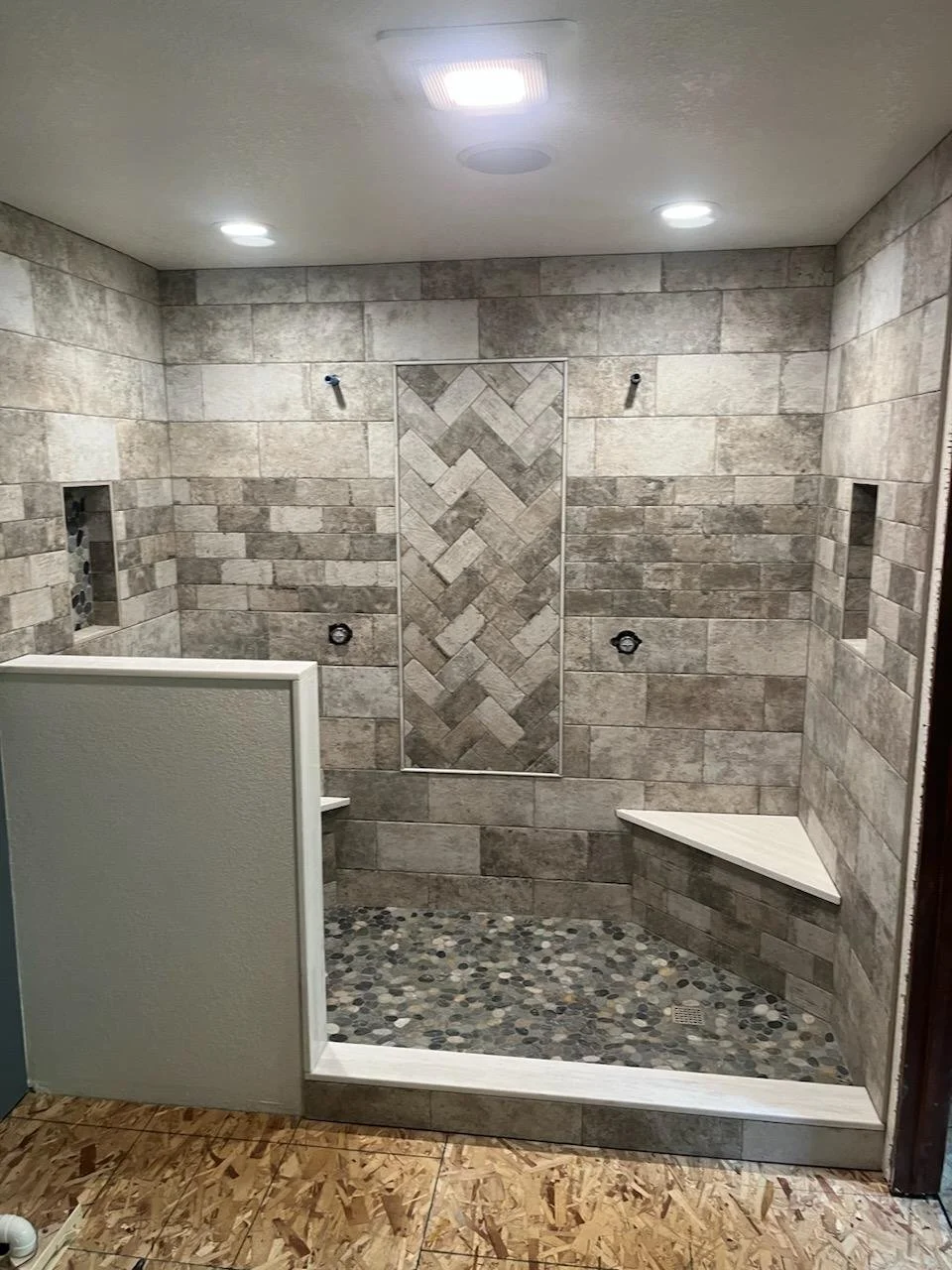 A partially finished walk-in shower with tiled walls, pebble stone floor, niche shelves, and a corner bench, under bright ceiling lights.