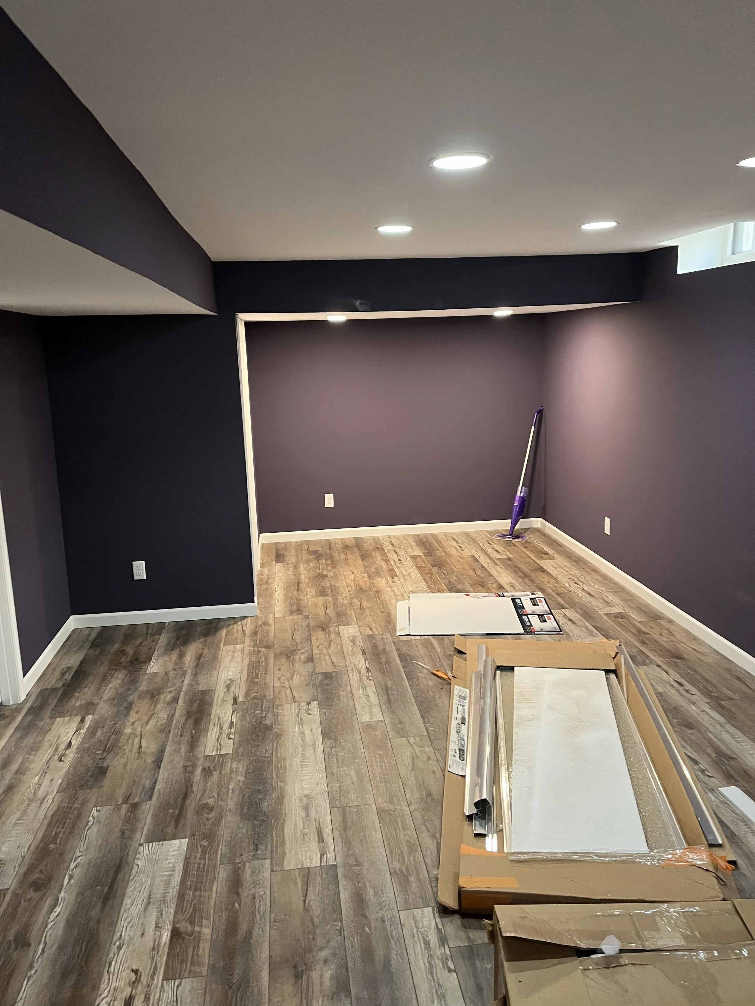 Empty basement room with purple walls, wood flooring, recessed ceiling lights, and partially assembled furniture or fixtures, with a vacuum cleaner in the corner.
