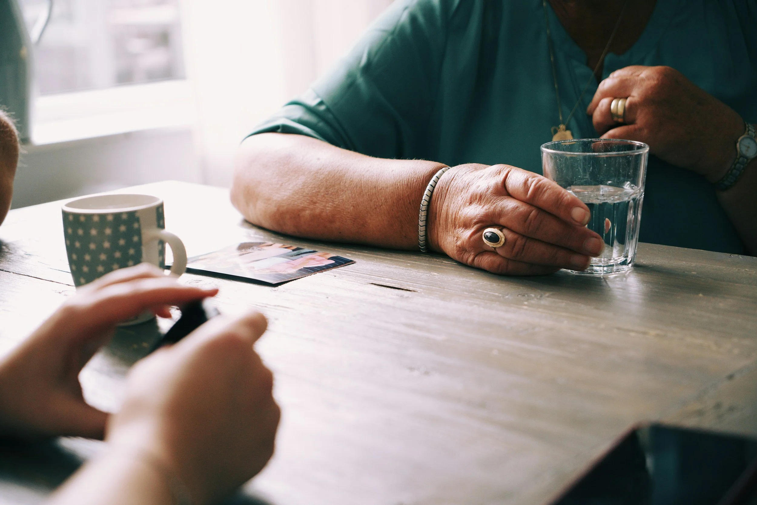 An elderly person with jewelry on their fingers and wrists is holding a glass of water. A mug with a star pattern and a photo are on the table, along with a child's hand holding a toy or object, and a window in the background.