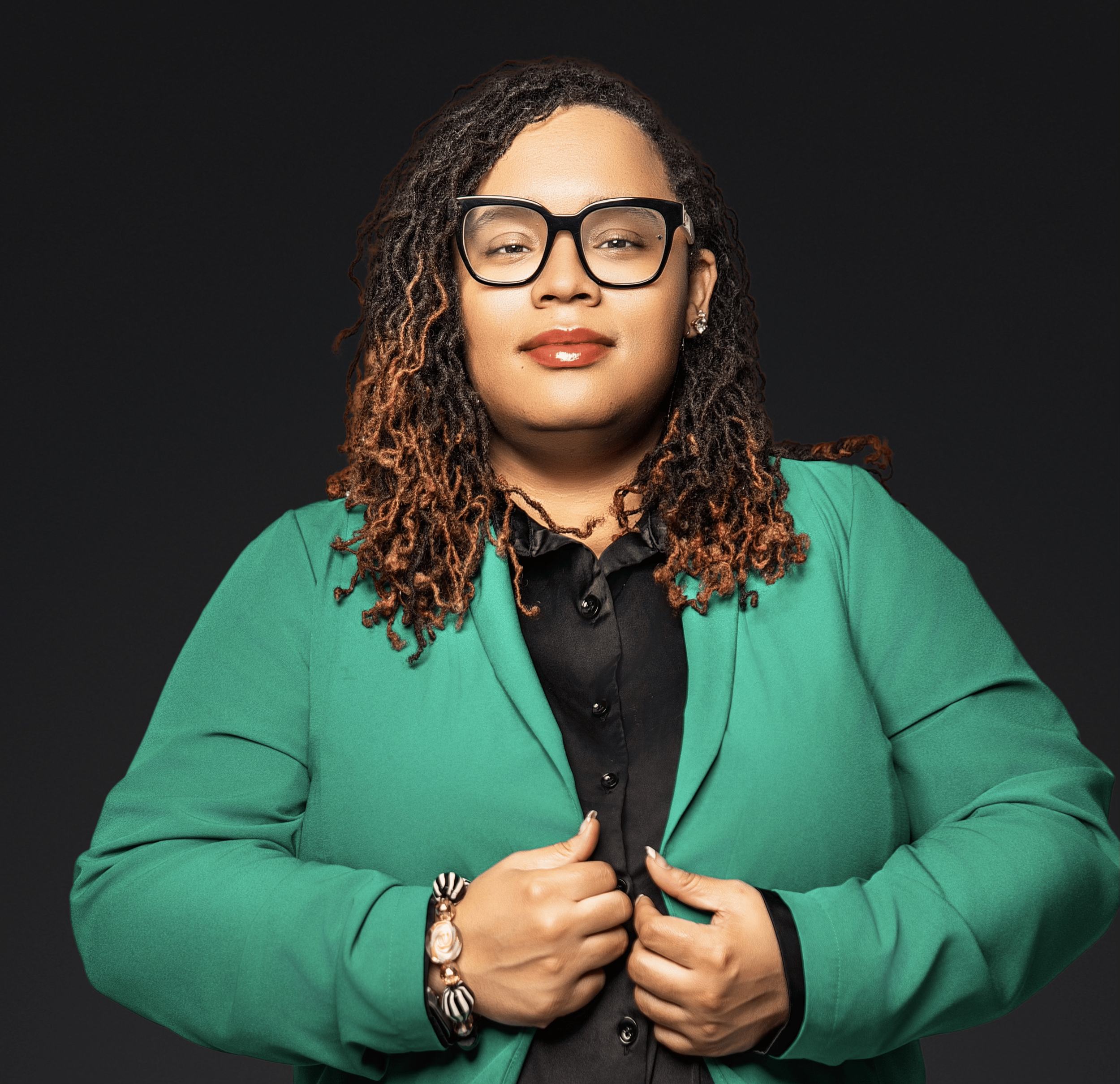 A woman with glasses, curly hair, wearing a green blazer over a black shirt, posing with confidence against a dark background.