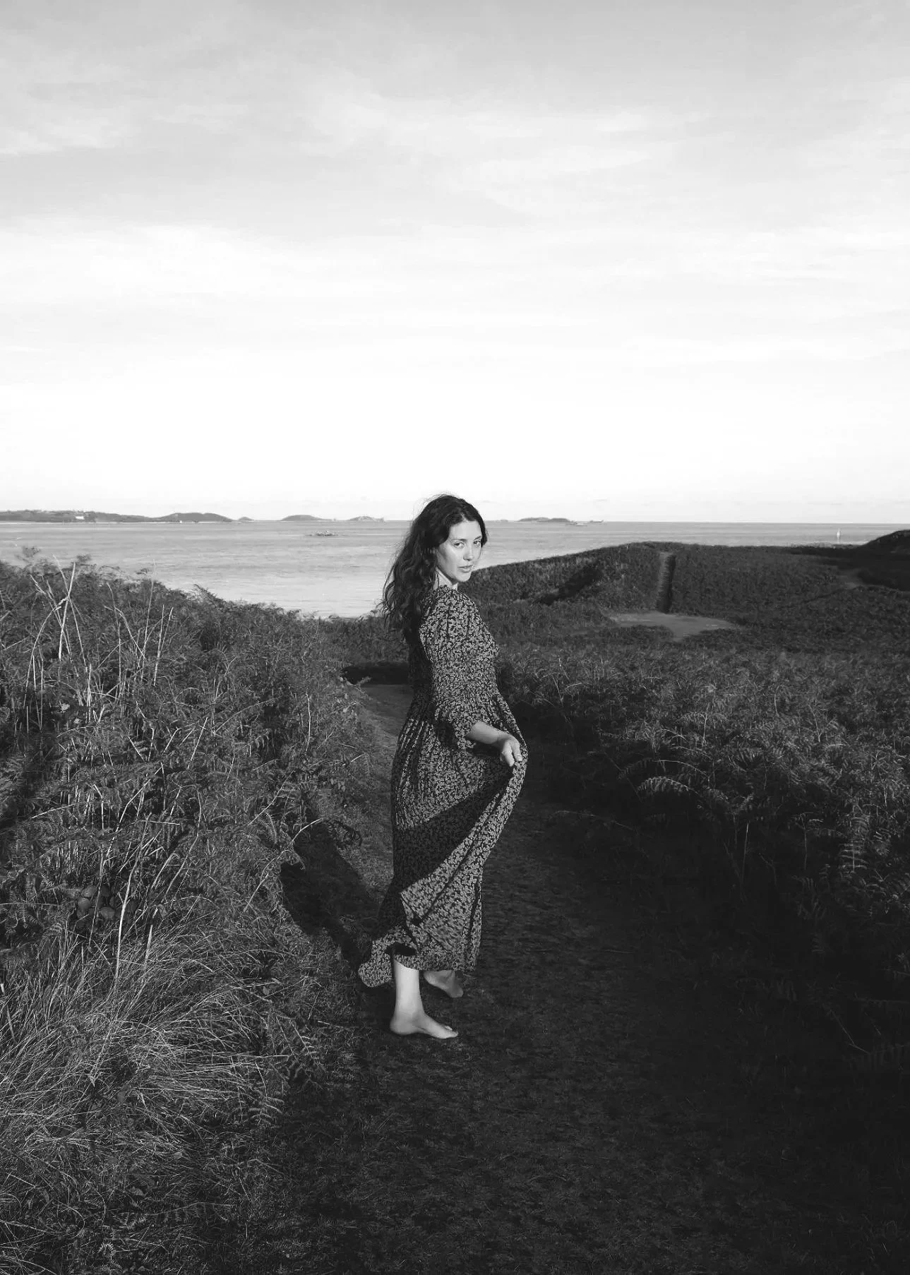 A woman standing barefoot on a dirt path near bushes on a coastline with water and small islands in the background, wearing a long, flowing dress, looking over her shoulder.