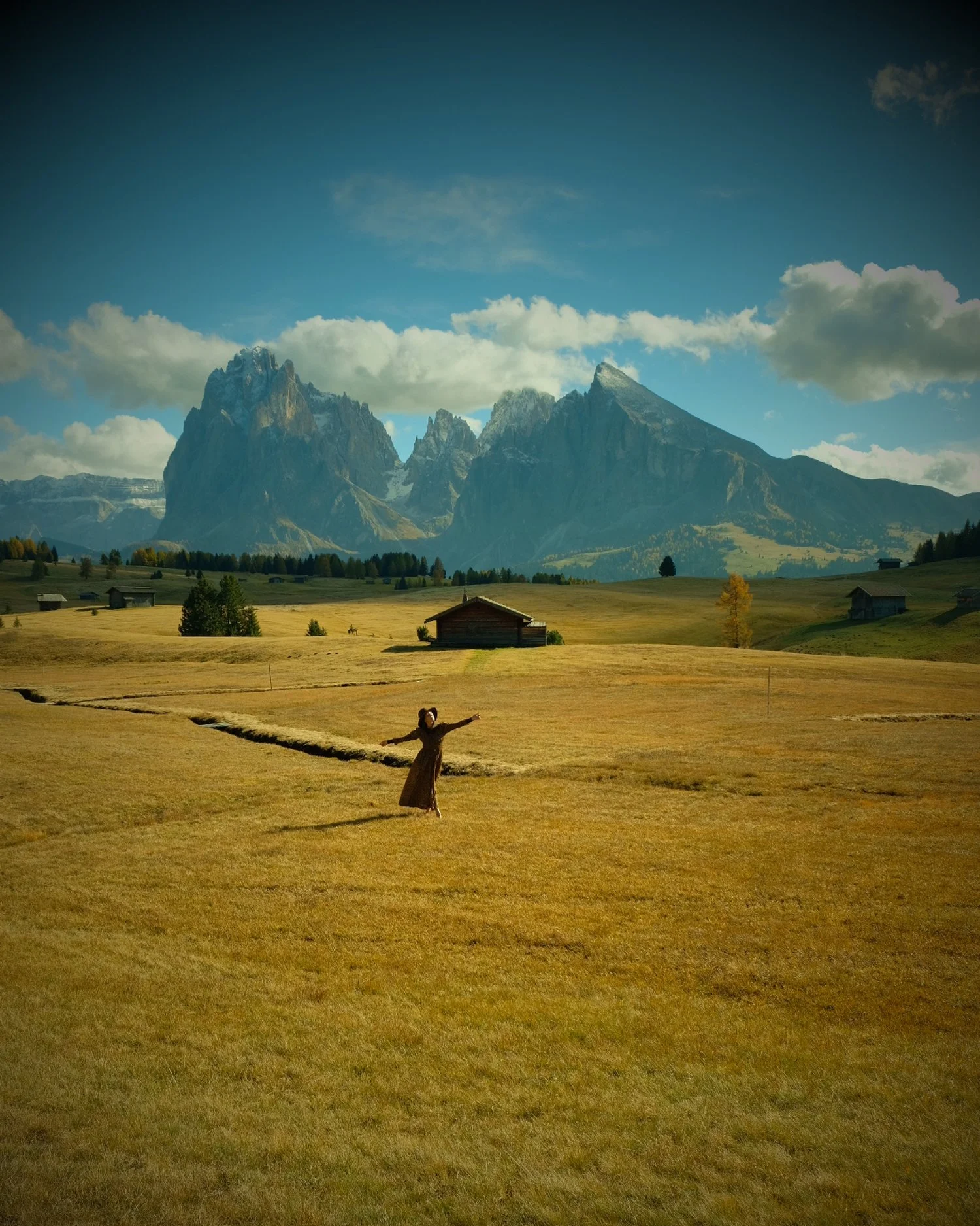 Woman in a brown dress standing with arms outstretched in a vast yellow field with mountains in the background under a partly cloudy sky.