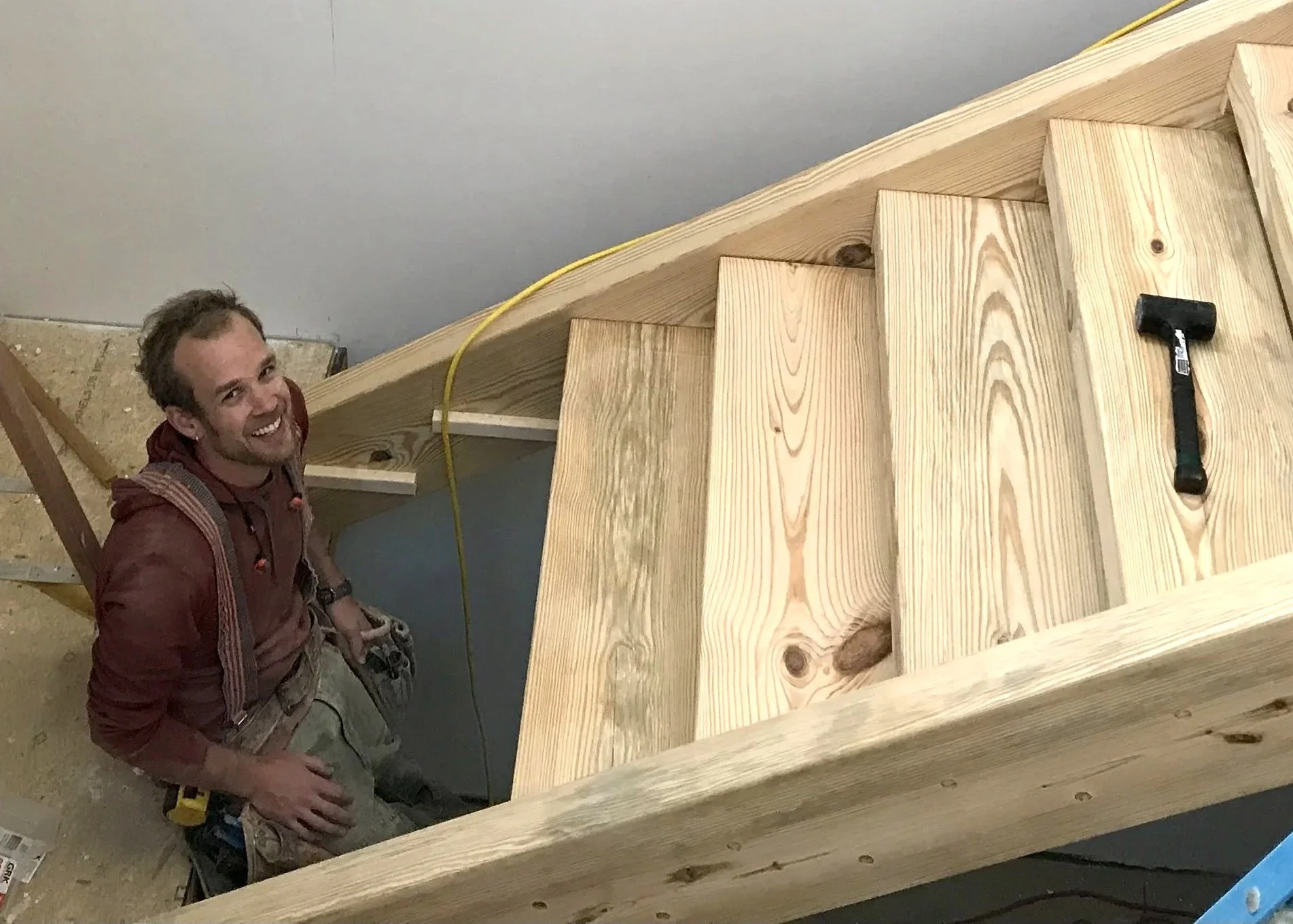 Kyle Zimmerman, a project coordinator and tradesman smiling inside a wooden staircase under construction, with tools nearby.