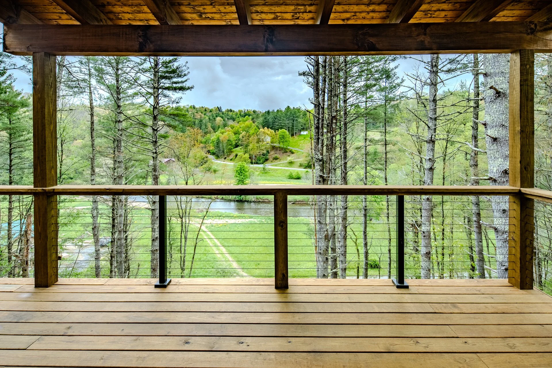 View from a wooden balcony overlooking a lush green landscape with trees, a winding road, a river, and distant hills under a cloudy sky. One of Sunny Day Homes' custom home builds on the New River in North Carolina.