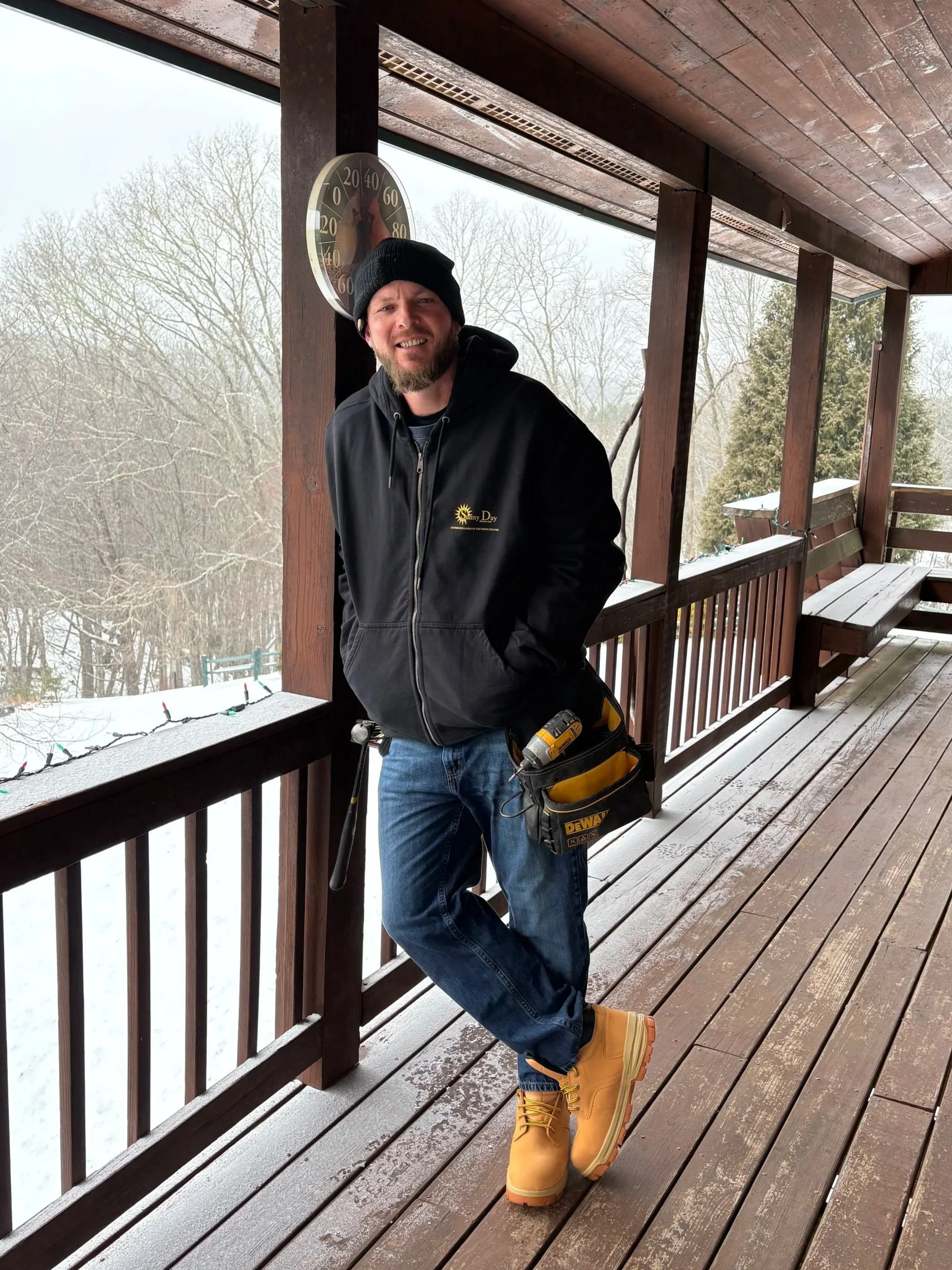 Phillip Mitchell Jr., A man with a beard and tattoos is taking a selfie outdoors with trees and a mountain in the background. He is wearing a gray striped polo shirt with a patch of a man with a hat and smoking a pipe, and a gray baseball cap.