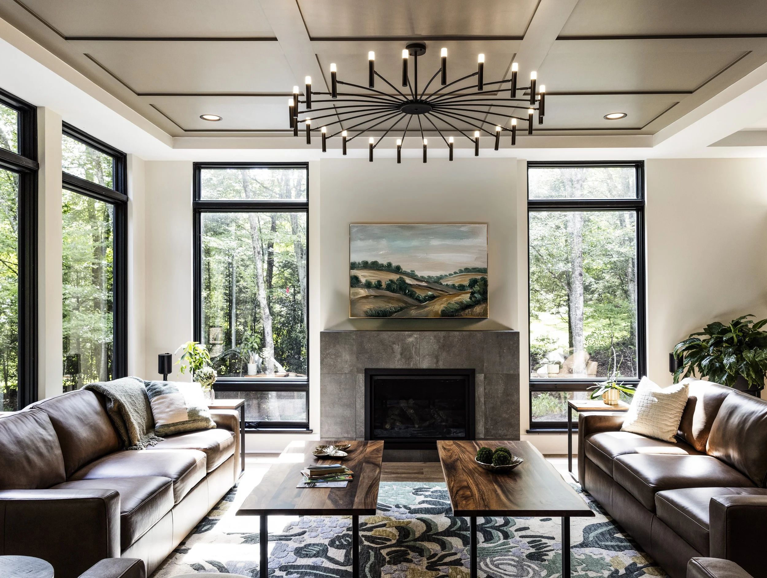 Living room with two leather sofas, wooden coffee tables, a modern chandelier, a fireplace with a landscape painting above it, and large windows showing greenery outside. One of Sunny Day Homes' custom home builds in Blowing Rock, NC.