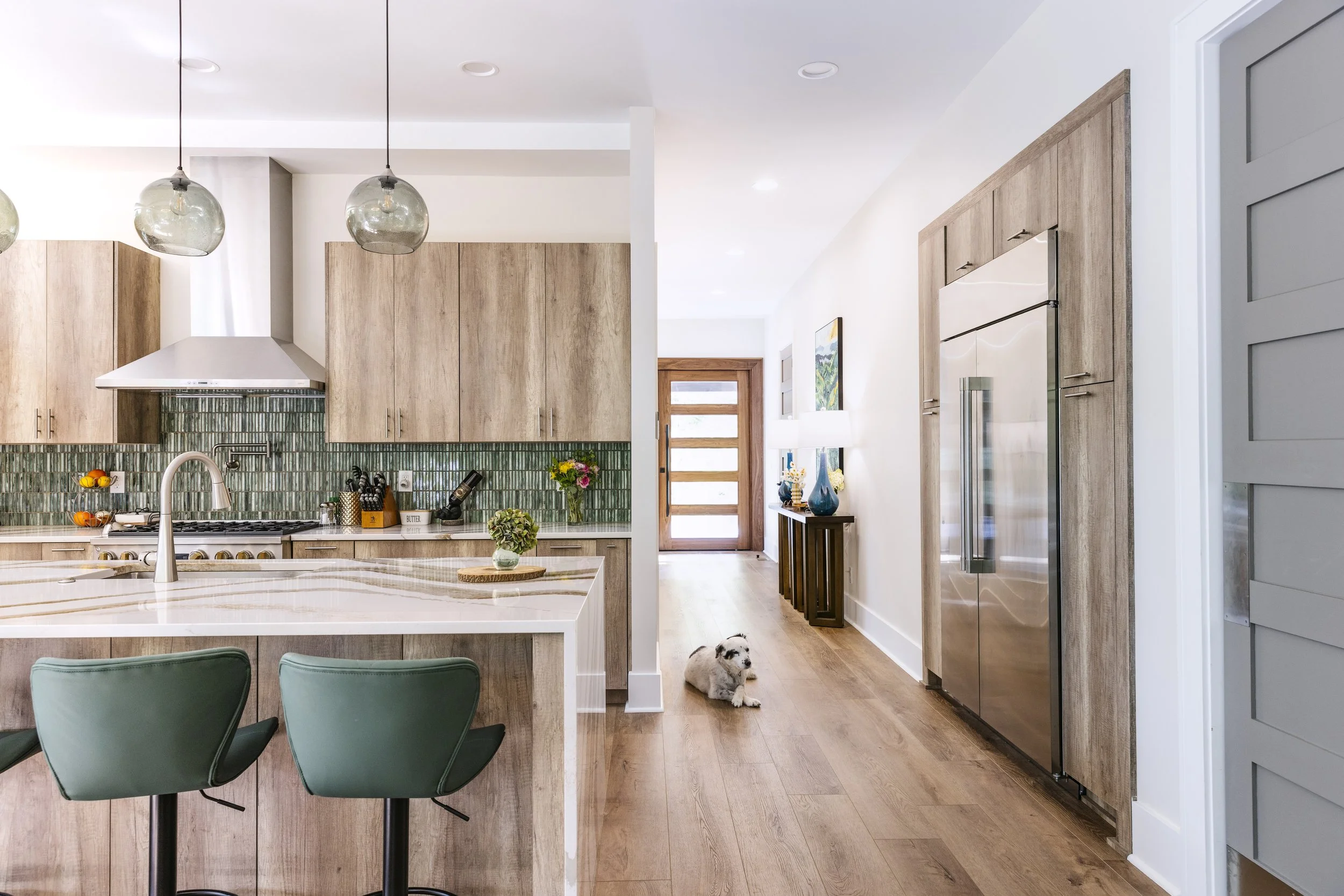 Modern kitchen with wooden cabinets, green backsplash tiles, a white island with two green bar stools, and a dog lying on the wooden floor in front of a wooden door. One of Sunny Day Homes' custom home builds in Blowing Rock, NC.