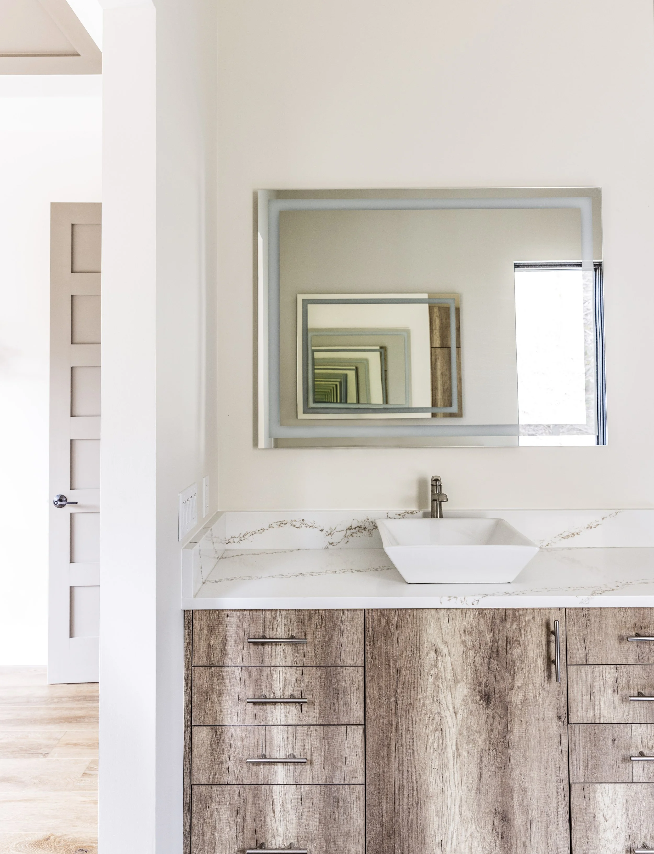 Modern bathroom vanity with a rectangular vessel sink, wooden cabinet with horizontal handles, and a framed mirror reflecting a series of smaller mirrors, a window, and part of a white wall. One of Sunny Day Homes' custom home builds in Blowing Rock,