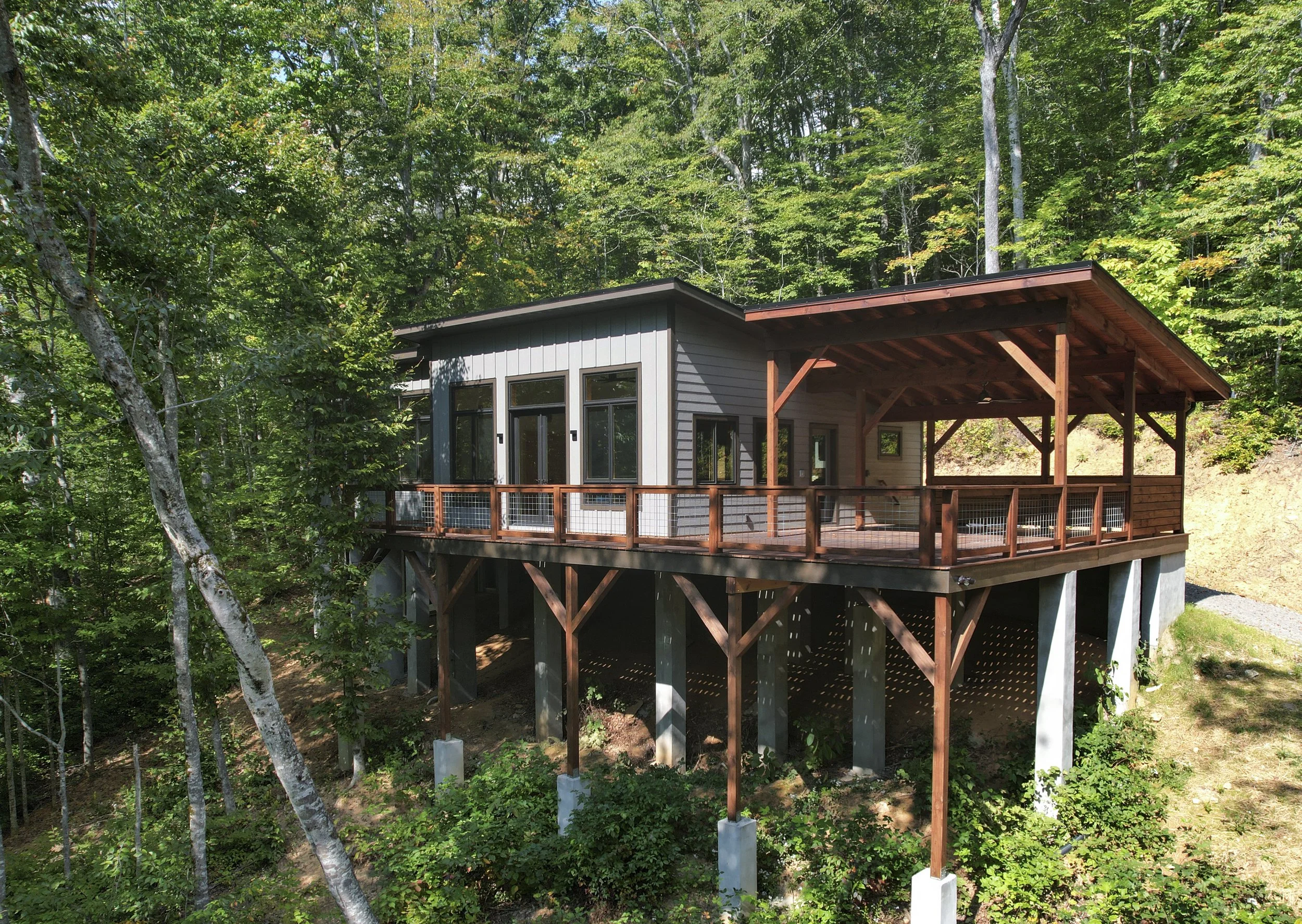 A modern house elevated on stilts with a large wooden deck, surrounded by green trees on a hillside. One of Sunny Day Homes' custom home builds in the High Country of NC.