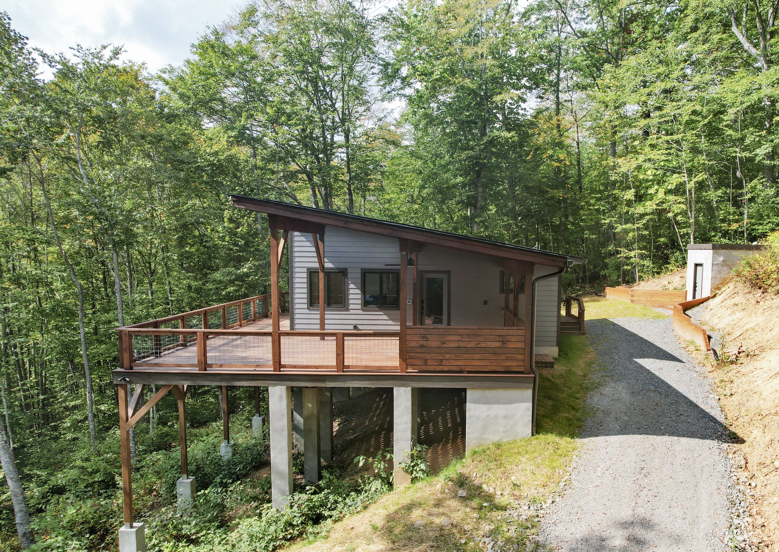 A modern house on stilts surrounded by green trees, with a large wooden deck and a gravel driveway. One of Sunny Day Homes' custom home builds on the New River in North Carolina.