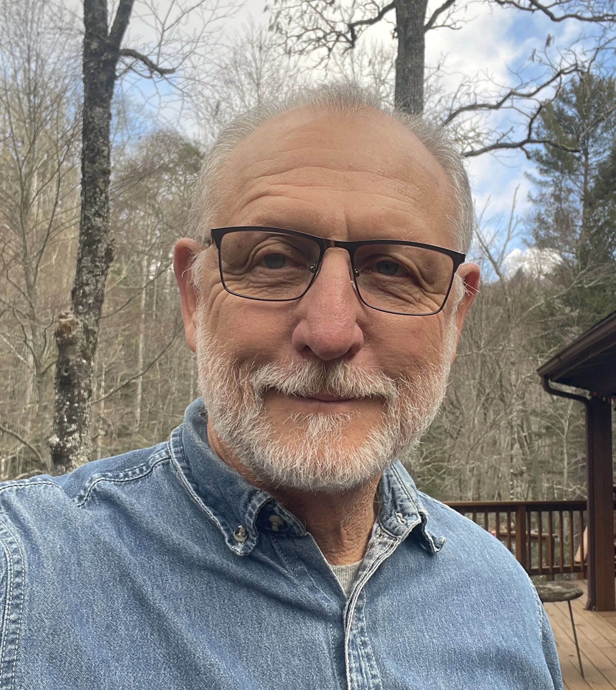 Sam Zimmerman, a man with glasses and a beard, smiling outdoors on a wooden deck with trees and a cloudy sky in the background.
