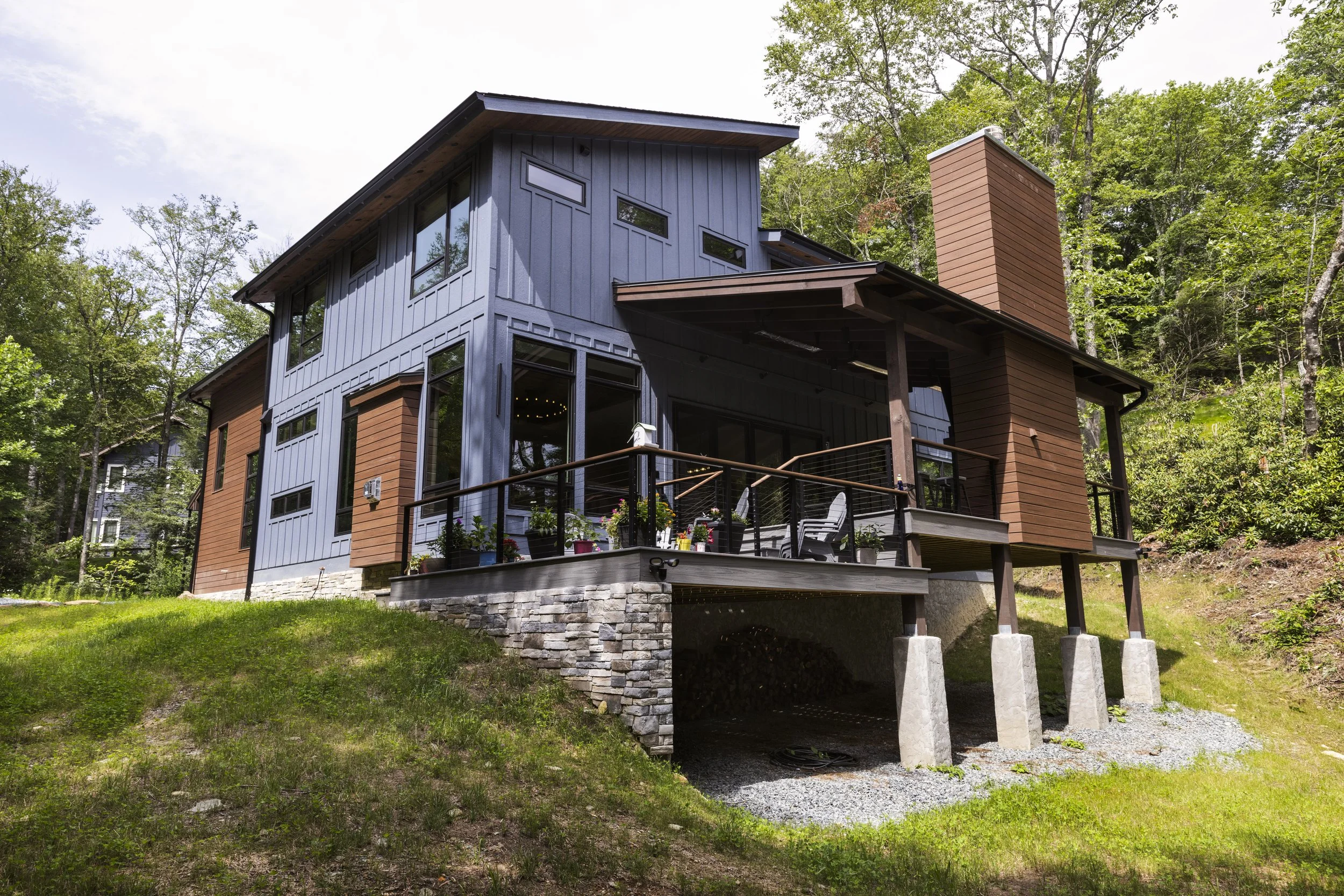 Modern house with blue and brown exterior, large windows, and a stone foundation, situated on a rhododendron filled hillside surrounded by trees. One of Sunny Day Homes' custom home builds in Blowing Rock, NC.