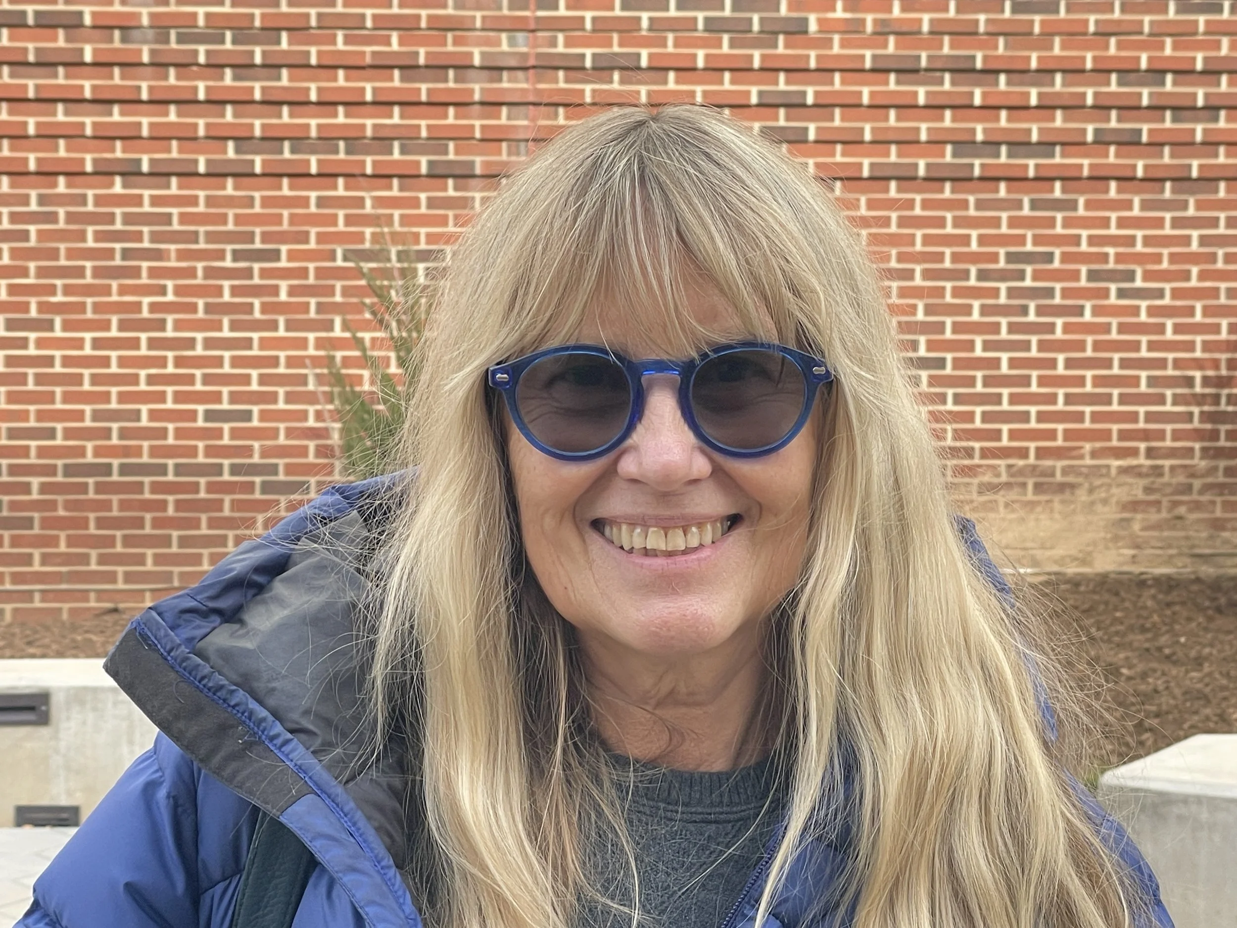 Joan Zimmerman, a woman with long blonde hair wearing blue glasses and a patterned knit sweater, smiling outdoors in sunlight.