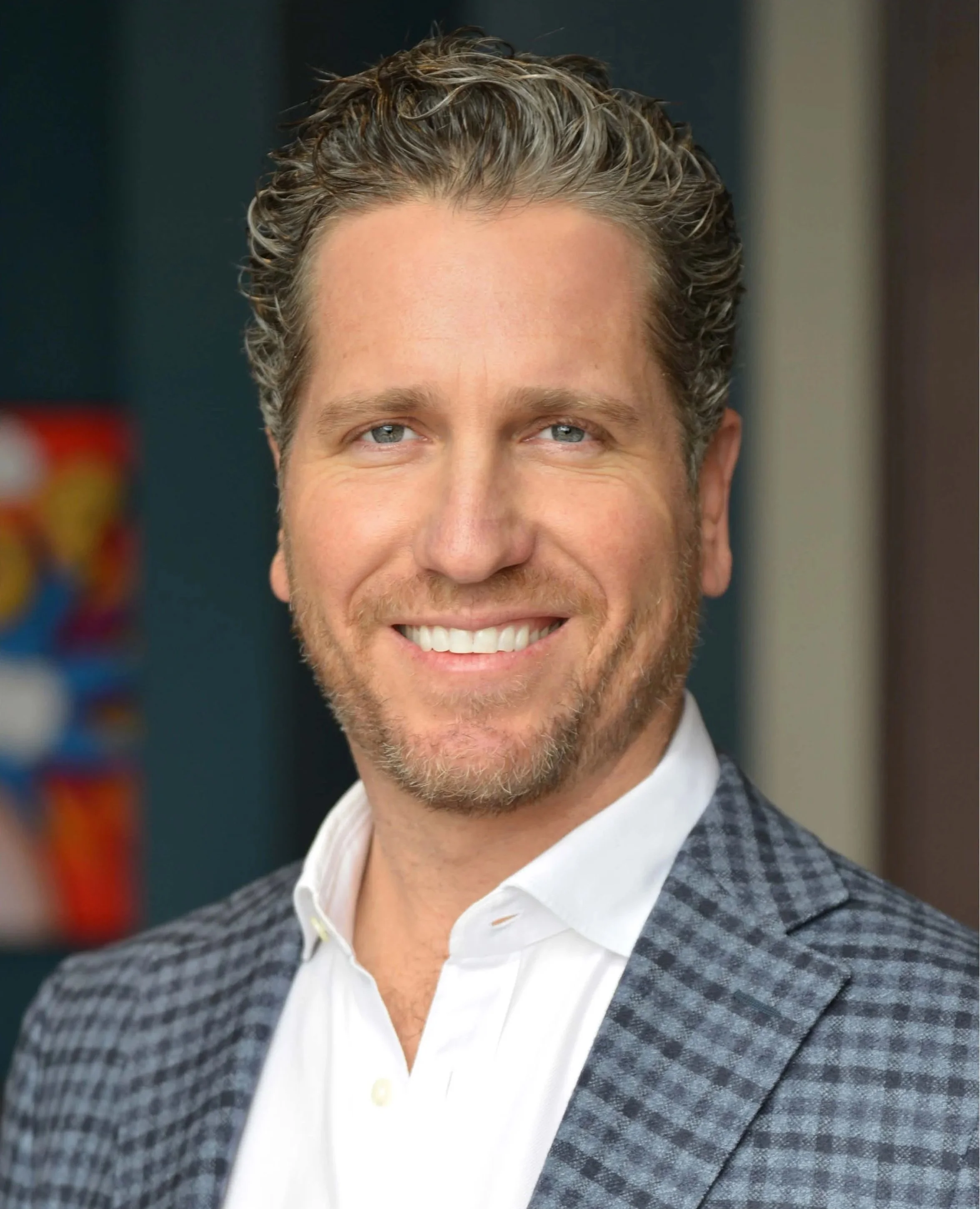 Headshot of smiling man with light brown hair, blue eyes, dressed in a white shirt and a checkered suit jacket.