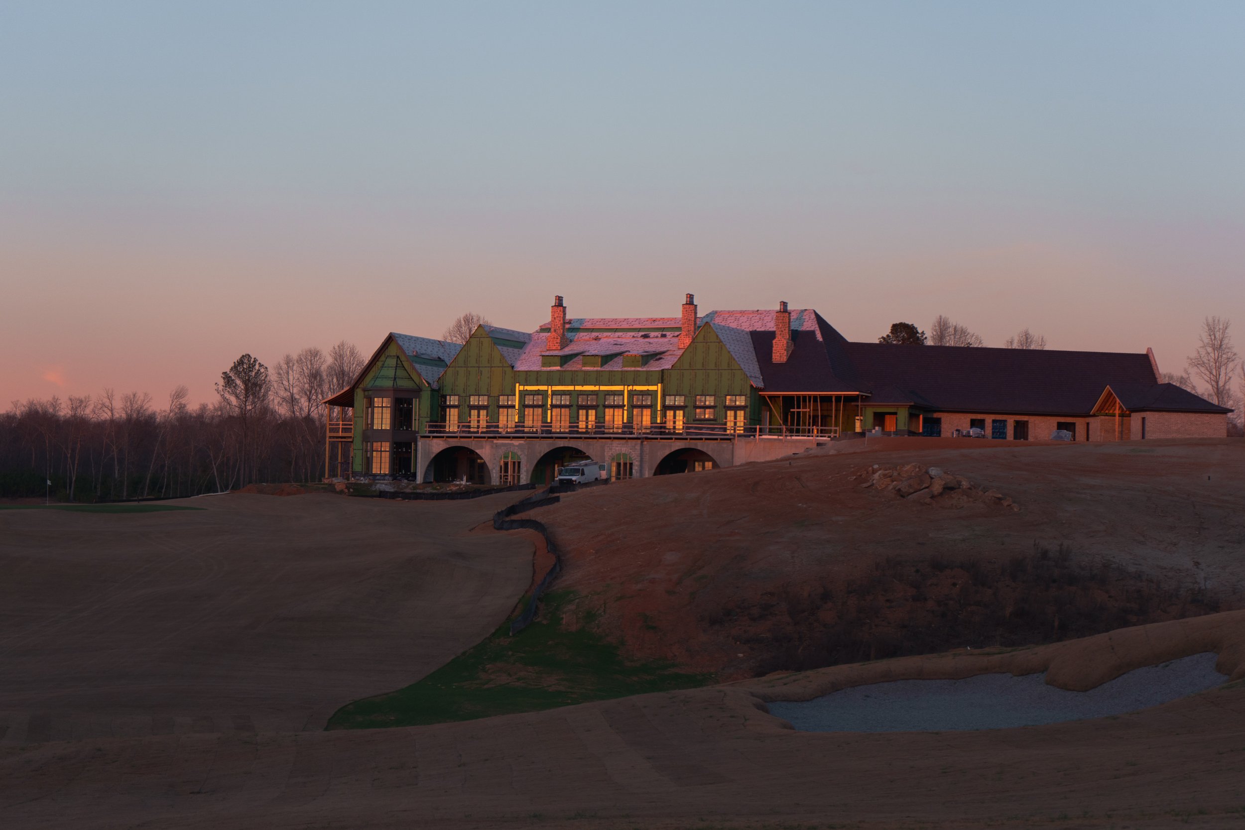 Construction site of a large house with green exterior walls, multiple chimneys, and arched doorways, situated on a hill at sunset.