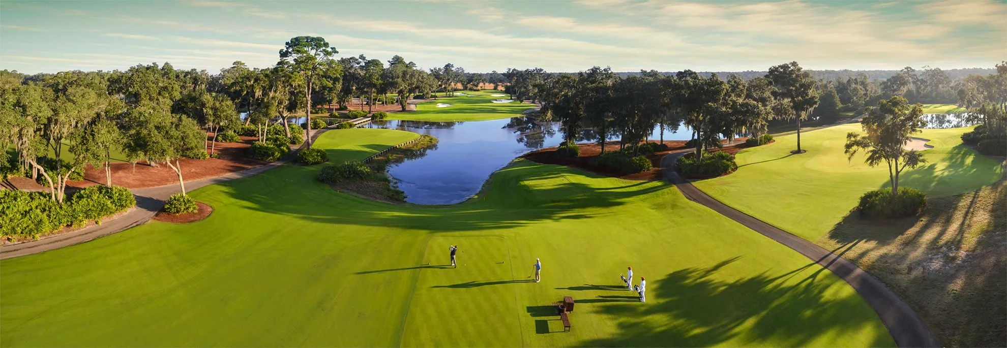 Aerial view of a golf course with green fairways, sand traps, water hazards, and tall trees, with a group of golfers on the putting green.