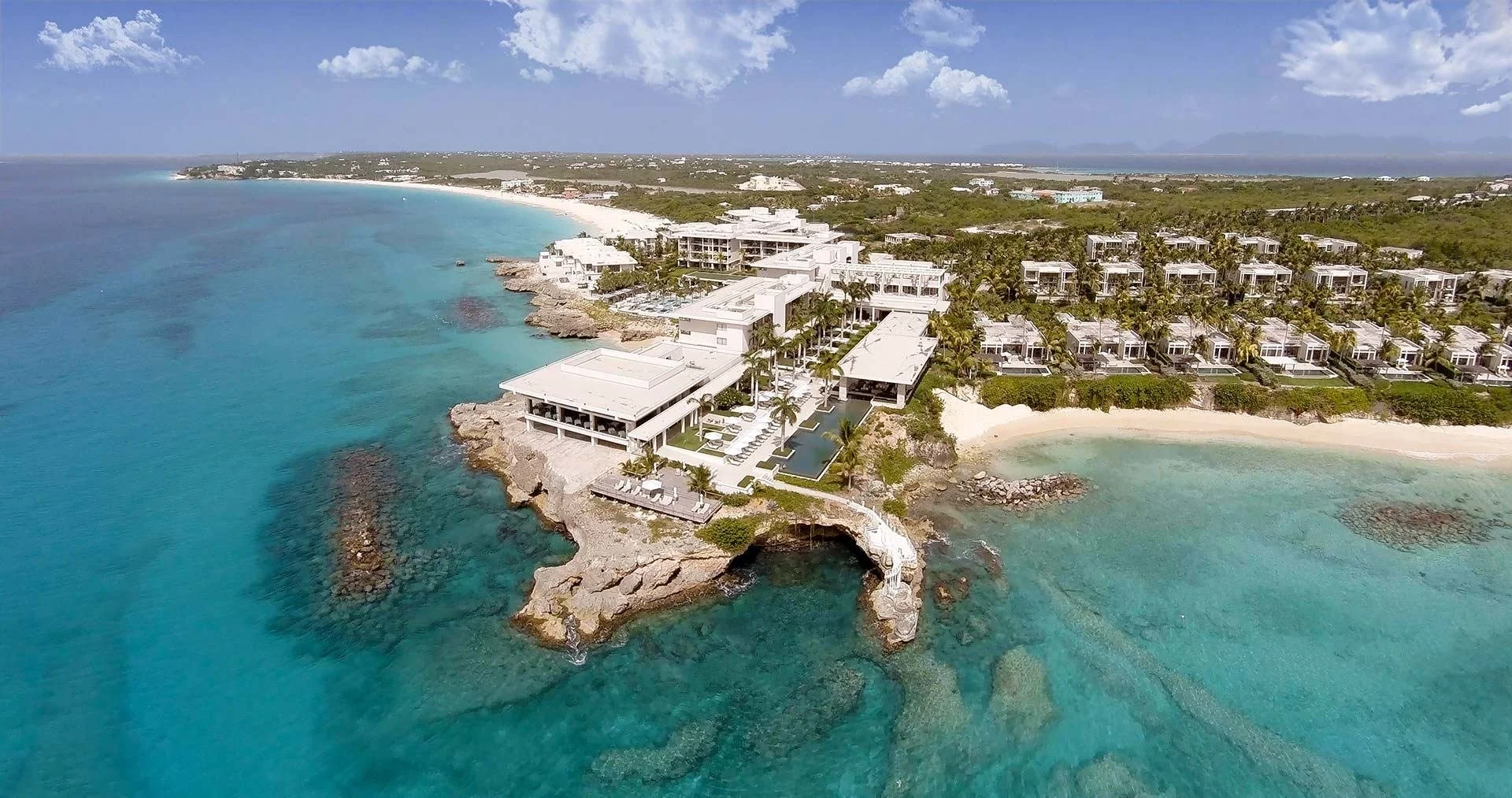 Aerial view of modern beachfront resort buildings surrounded by turquoise ocean water and sandy beaches with lush greenery in the background.