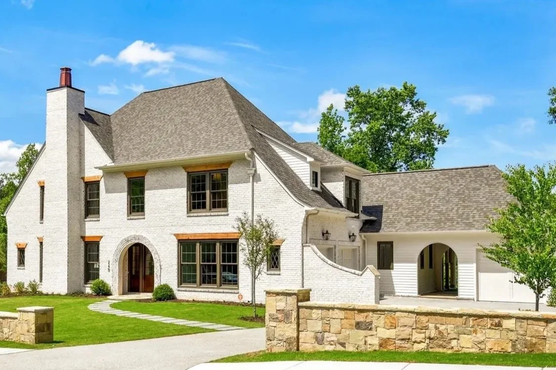 A white two-story house with a brown roof, stone accents, front porch, and arched entryway, surrounded by a green lawn and trees under a blue sky.