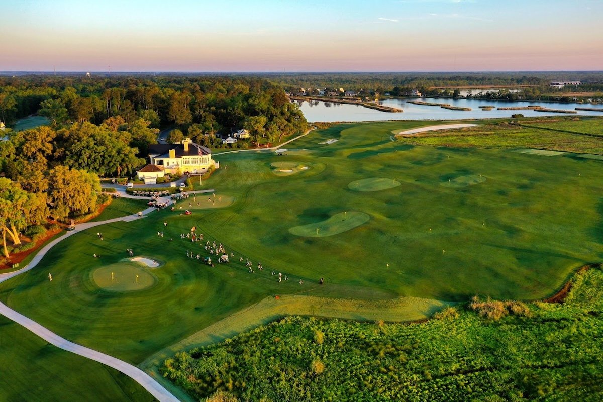 Aerial view of a golf course with sand traps, putting greens, carts, and a clubhouse near a water body, surrounded by trees during sunset.