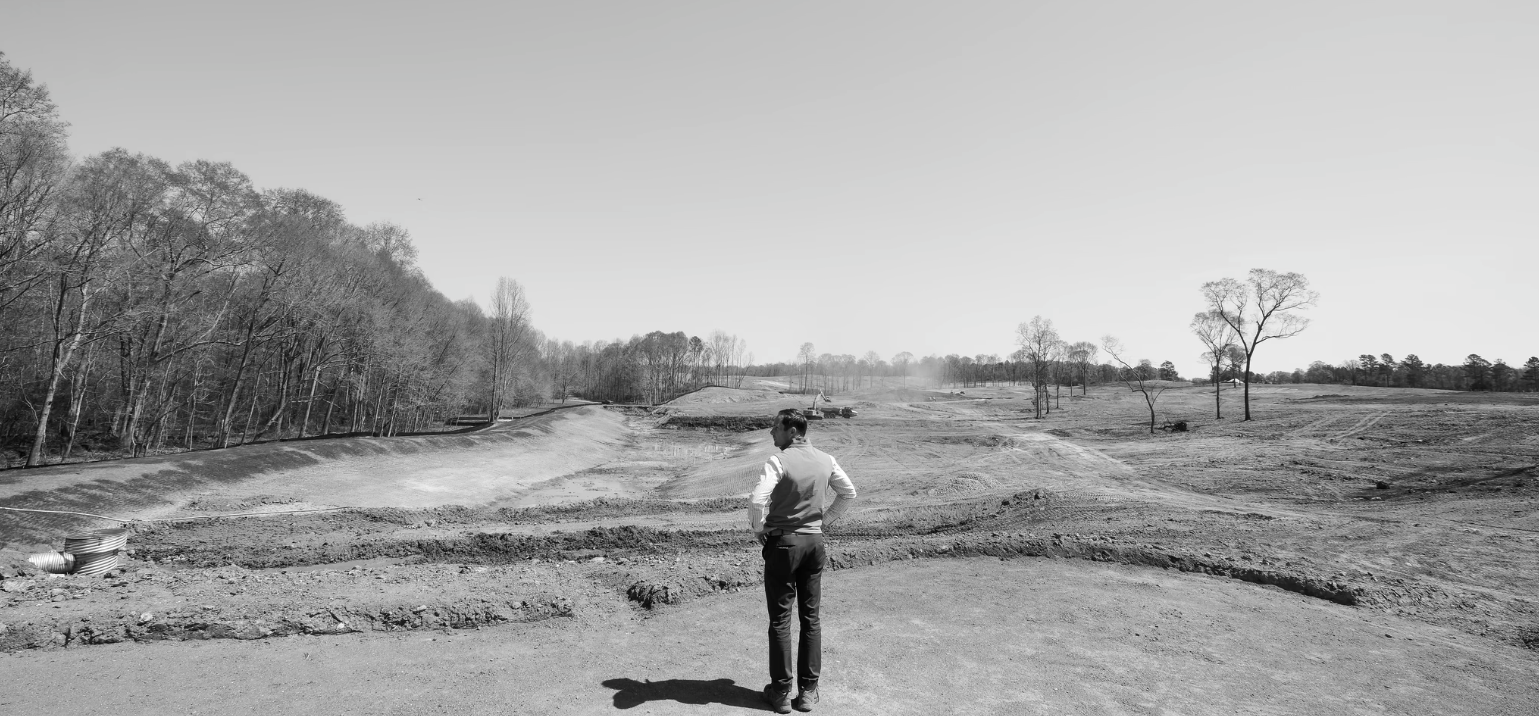 A man in business attire stands on a dirt construction site, looking across the open area with sparse trees and a distant forest on the horizon.