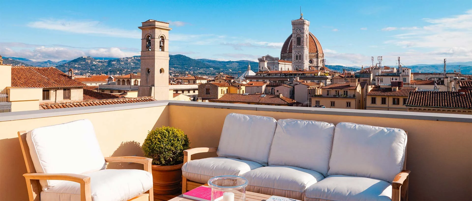 A rooftop terrace with white cushioned outdoor furniture, a potted plant, and a view of Florence, Italy, featuring the Florence Cathedral with its distinctive dome, and surrounding historic buildings under a clear blue sky.