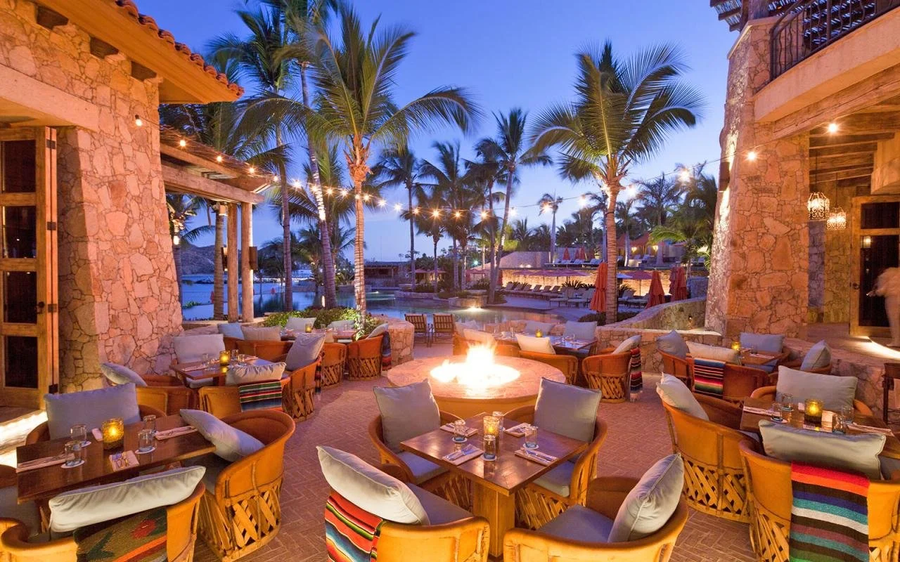 Outdoor patio at a resort during evening with a fire pit, palm trees, string lights, and tables set for dining.