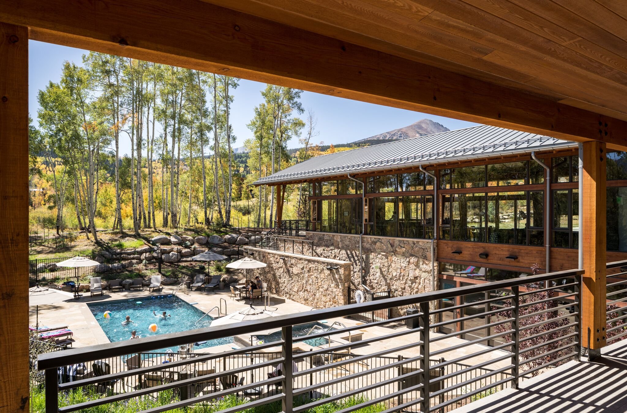 View from a balcony looking down at a swimming pool with people swimming, surrounded by lounge chairs and umbrellas, with a stone building, trees, and mountains in the background on a sunny day.