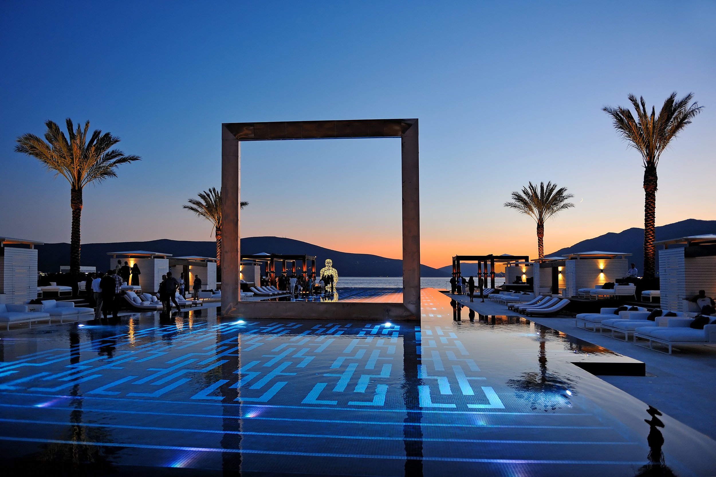 Luxury poolside lounge area at sunset with palm trees, modern cabanas, outdoor seating, and a view of mountains and the ocean in the background.