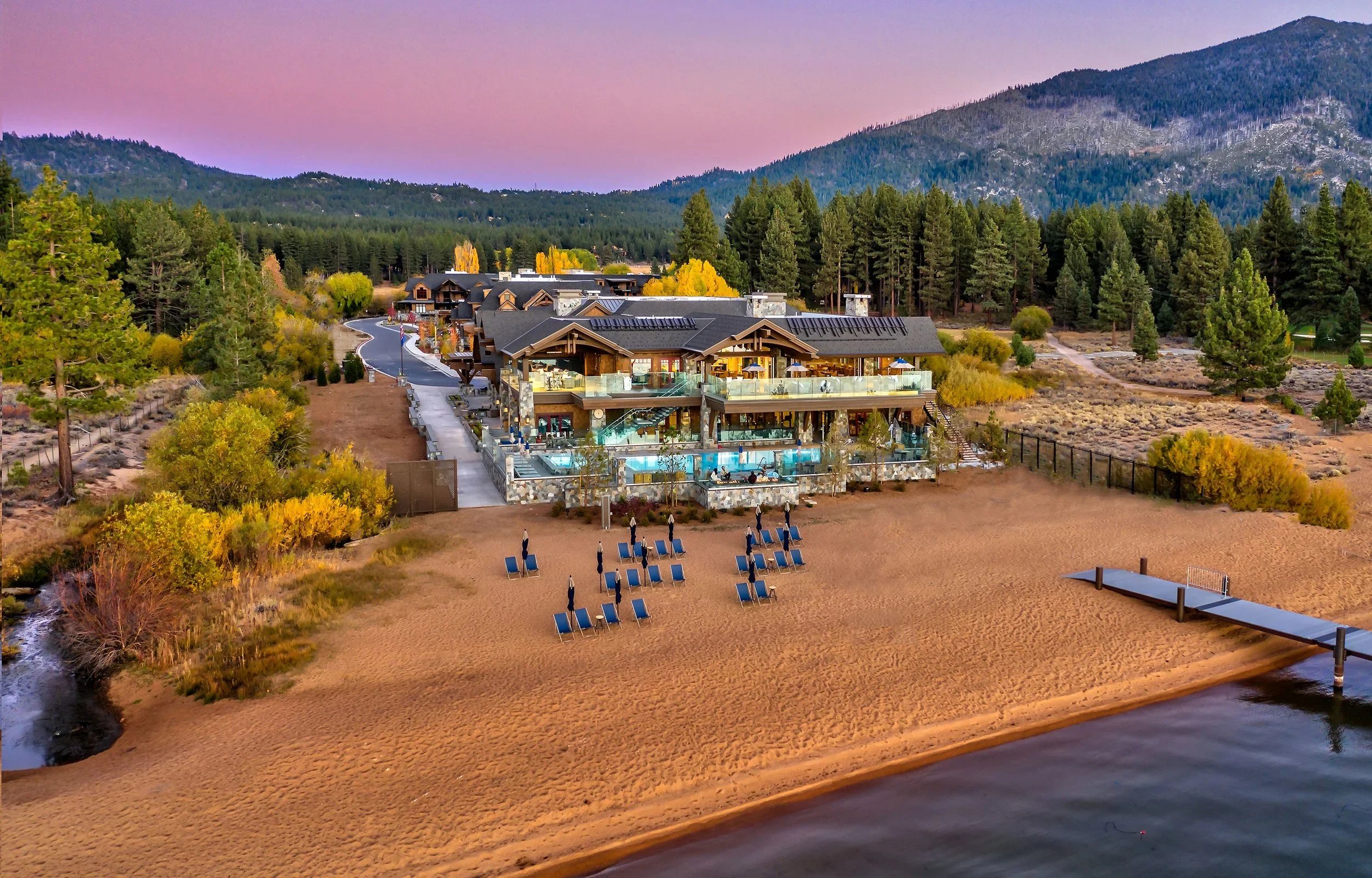 A lakeside resort with a large building featuring multiple balconies, an outdoor pool, and lounge chairs on a sandy beach, surrounded by trees and mountains at sunset.