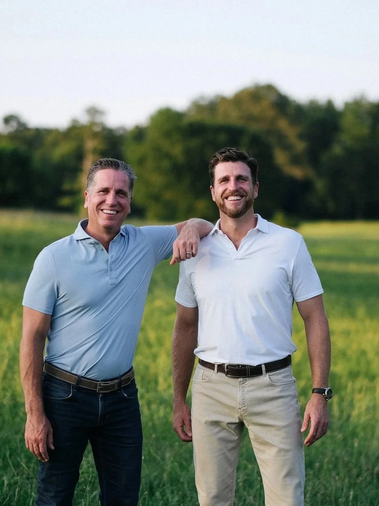 Two men standing outdoors on a grassy field with trees in the background, smiling at the camera. One man has his arm around the other's shoulder.