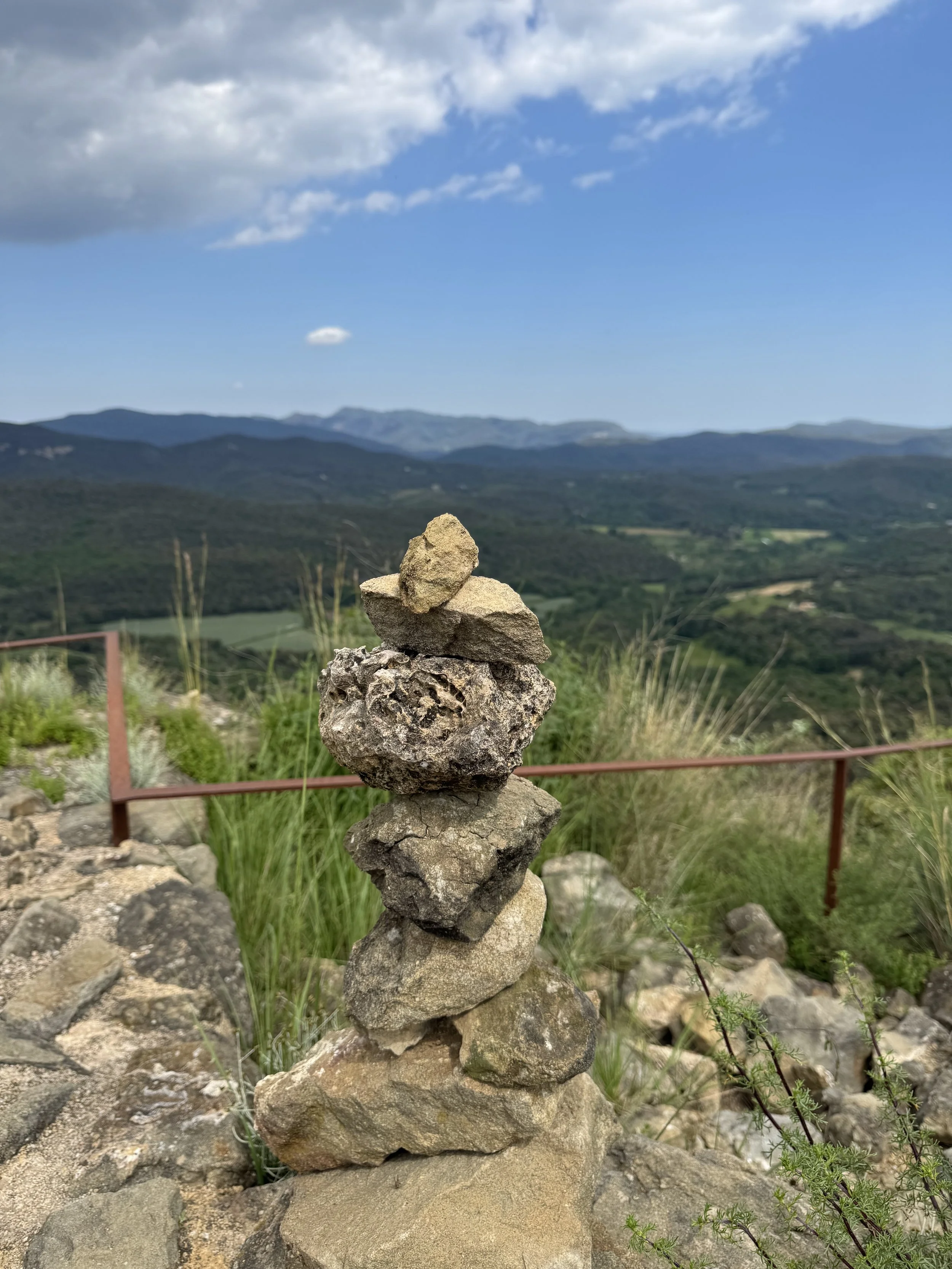 Stacked rocks on a hillside with a view of rolling mountains and a partly cloudy sky.