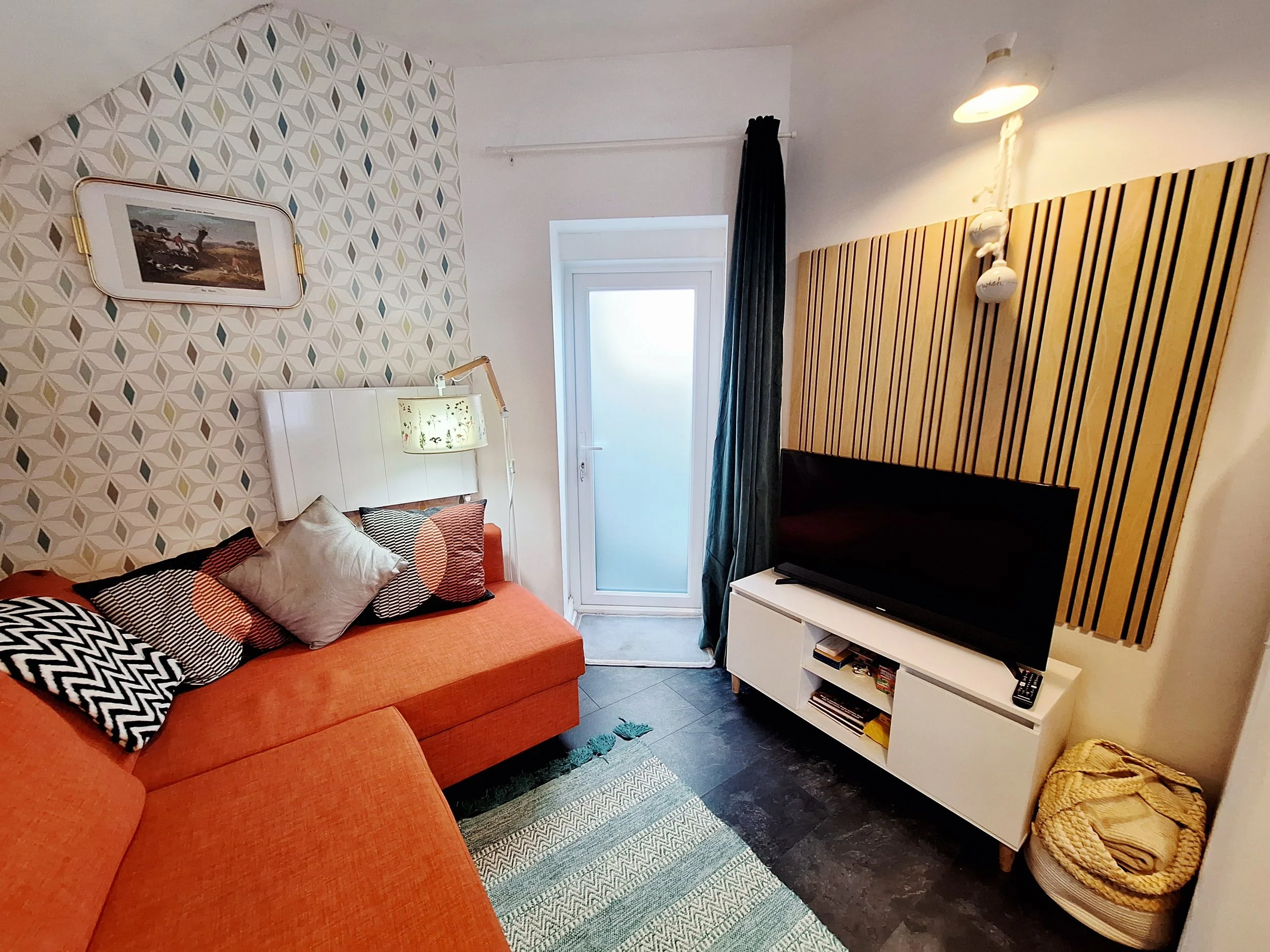 Living room with an orange sofa, patterned cushions, a wall-mounted light, a wall with geometric wallpaper, a television on a white stand, a window with a dark curtain, and a small basket with blankets.