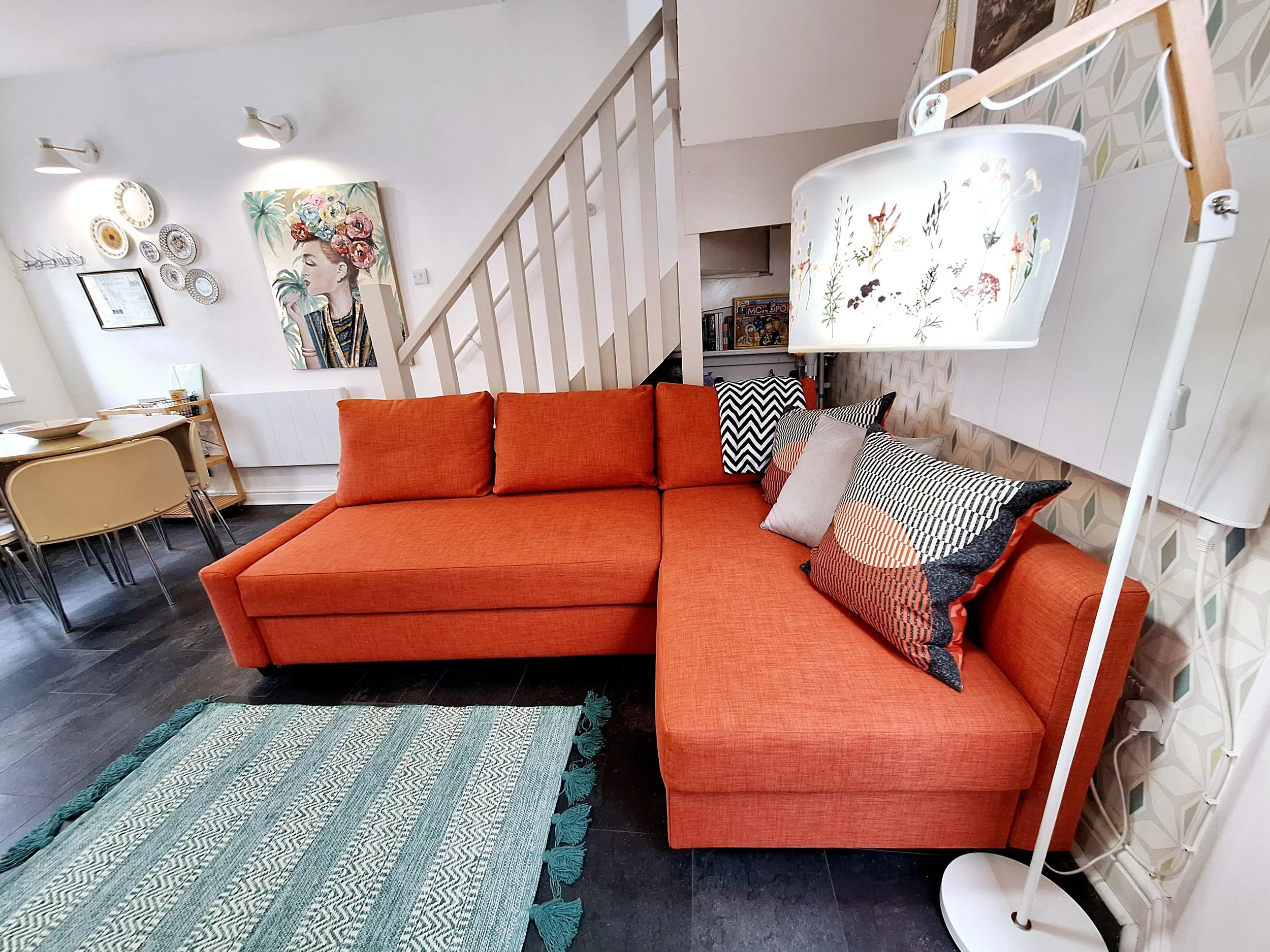 Living room with an orange sectional sofa, patterned pillows, a hanging lampshade with floral design, a blue rug, a dining area with a beige table and chairs, wall art including a portrait of a woman with flowers in her hair, and a staircase with a white railing.