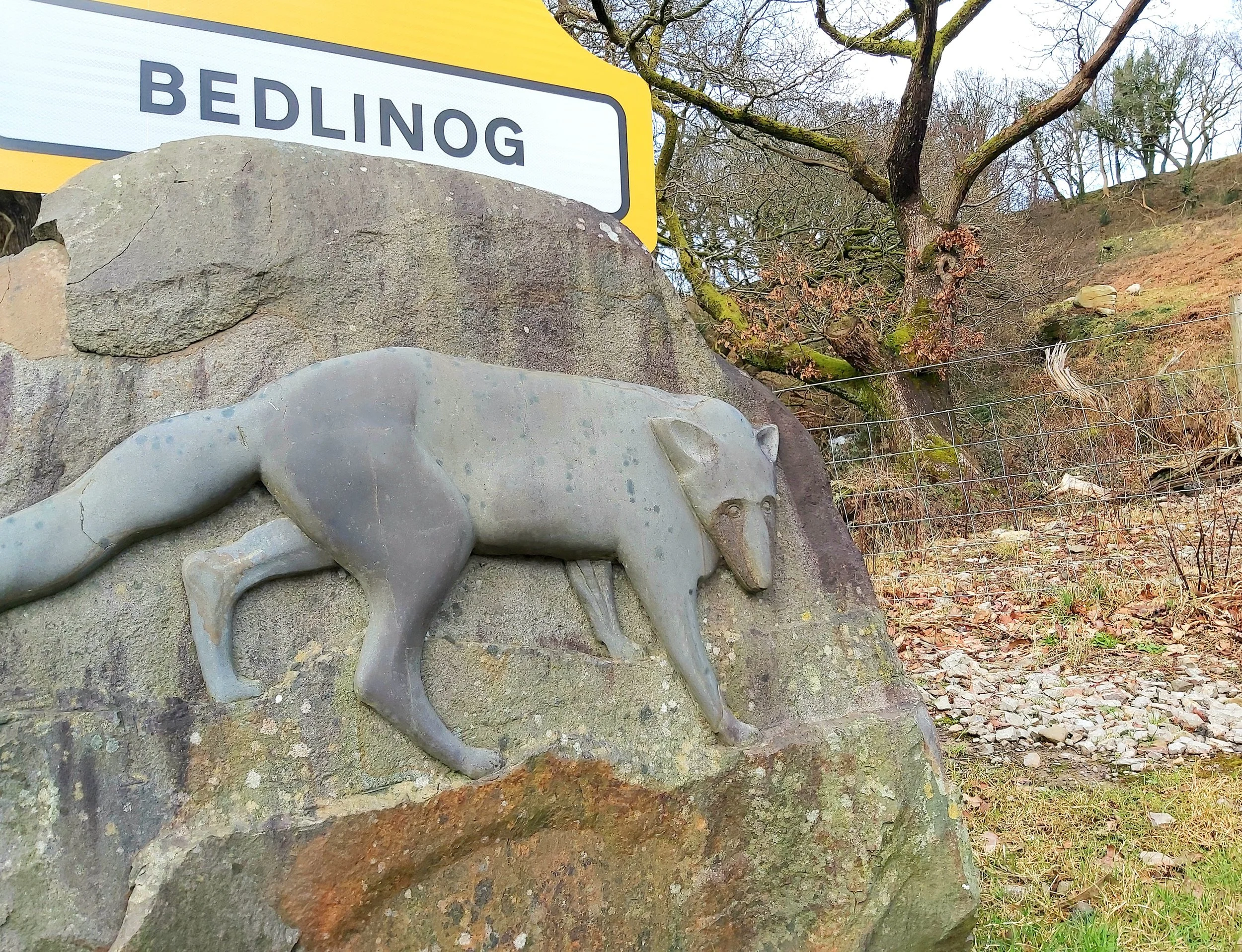 A stone sculpture of a wolf on a rock in a natural outdoor setting with a sign reading 'Bedling' and trees in the background.