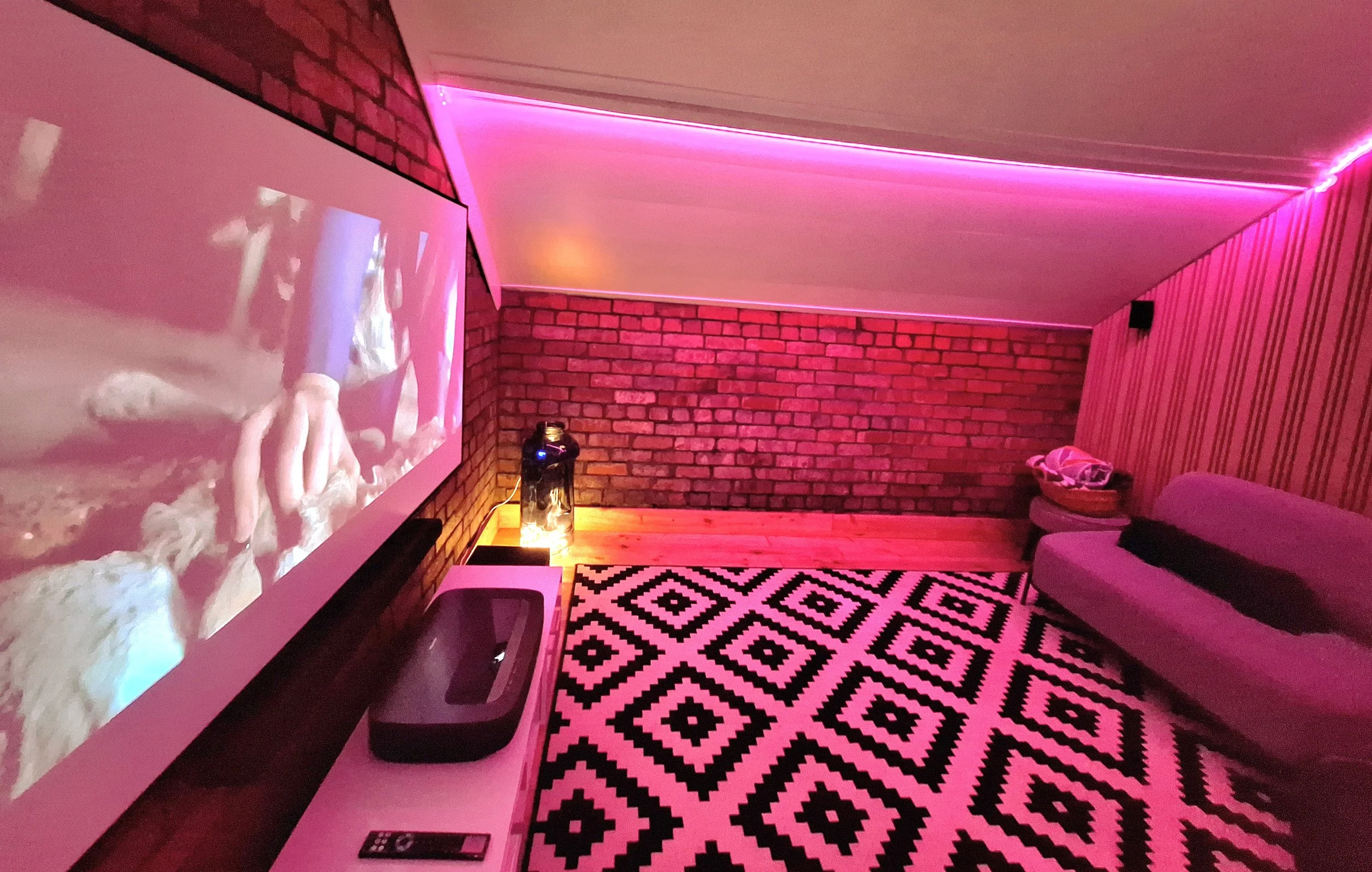 A cozy living room illuminated with pink and purple neon lights, featuring a brick wall, a patterned black and white rug, a sofa with a throw pillow, a side table with a basket, and a large wall-mounted screen showing a person knitting.