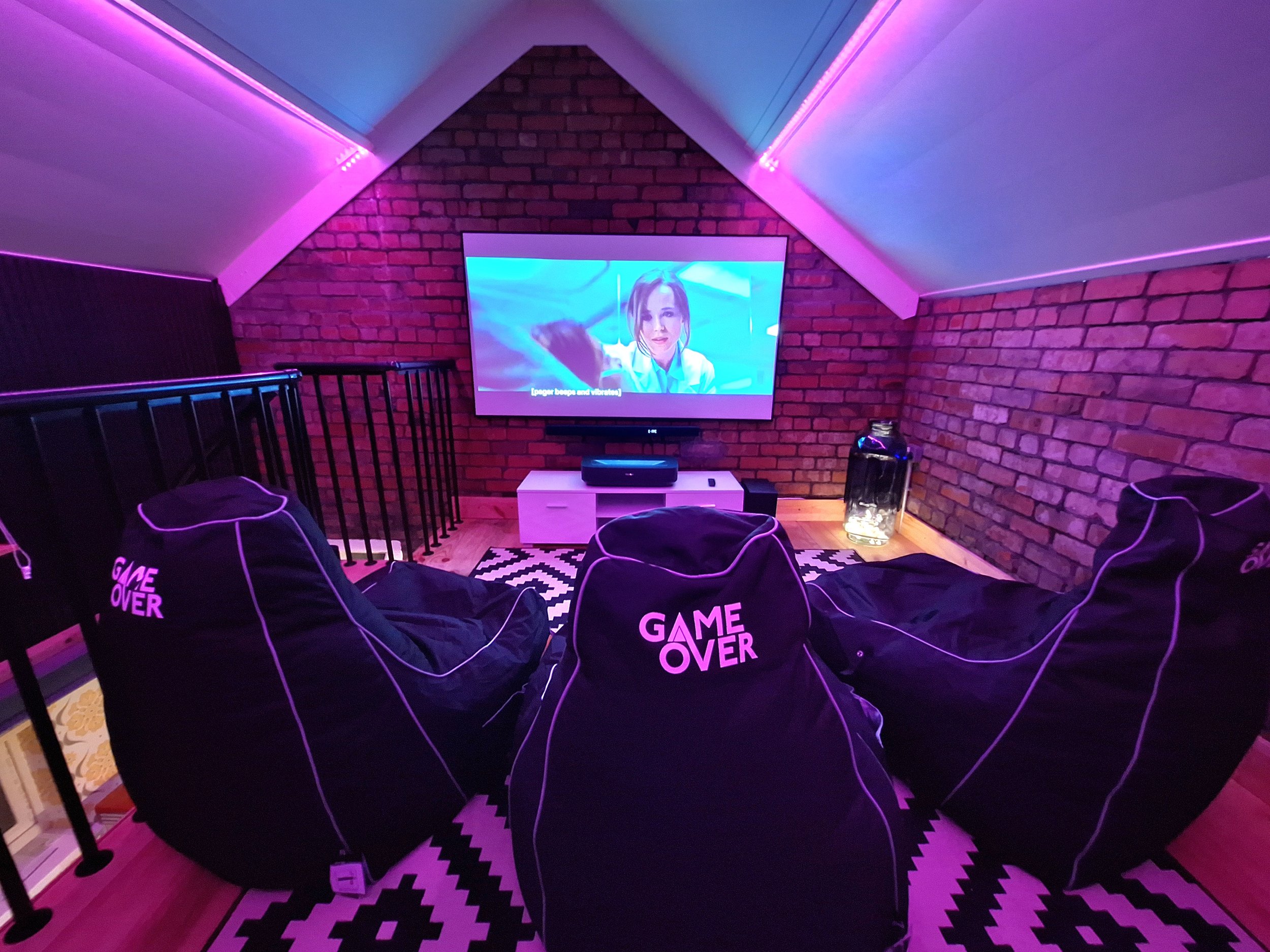 A cozy gaming room with three bean bag chairs labeled 'GAME OVER' in front of a large wall-mounted TV. The room has purple and pink LED lighting, exposed brick walls, and a black and white patterned rug. A small white entertainment stand and a fish tank are also visible.