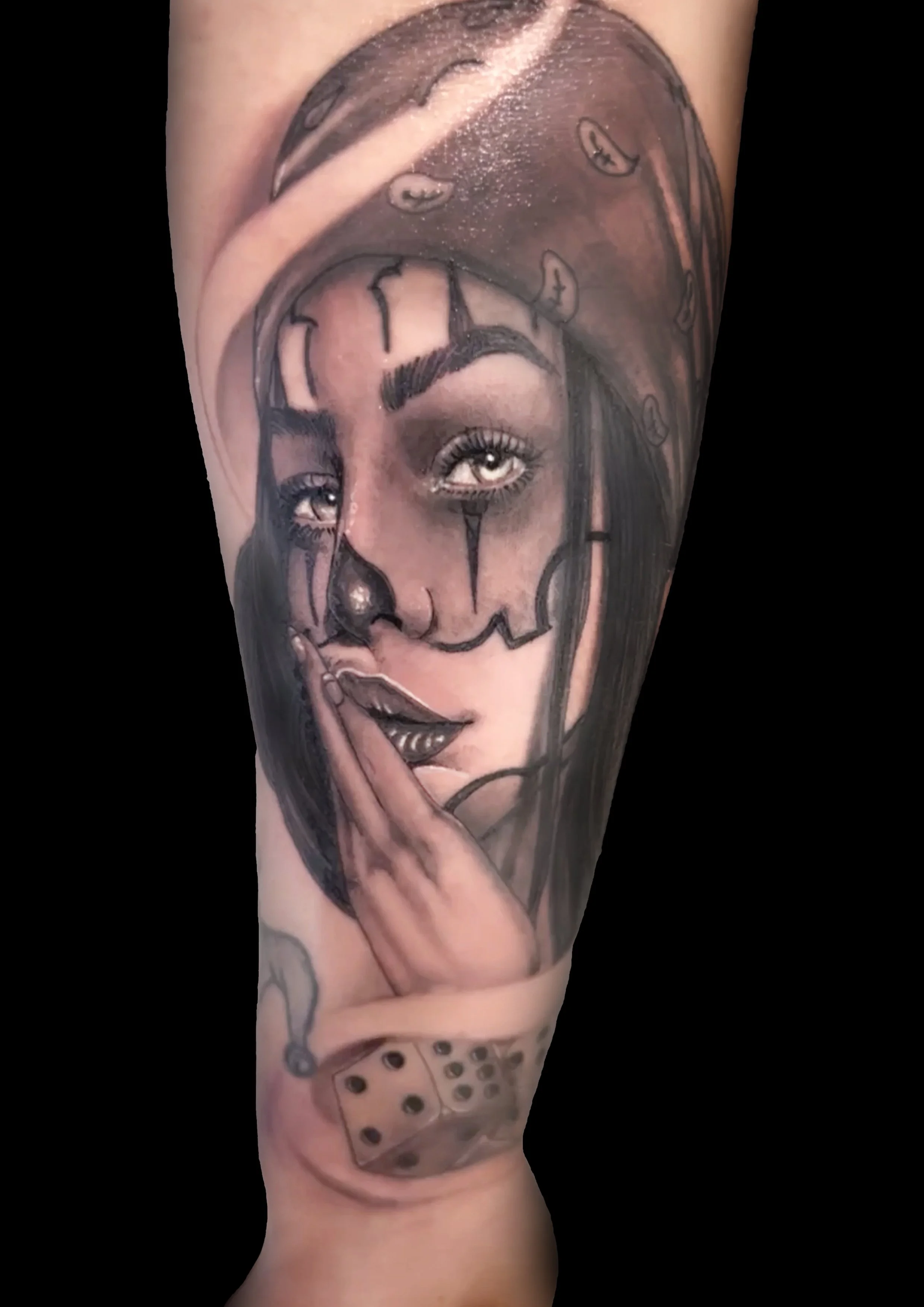 Tatuaggio realistico di una donna con una fronte da clown, occhi azzurri, labbra scure e una mano vicino al volto, con dettagli di orologio, dadi e altri elementi.