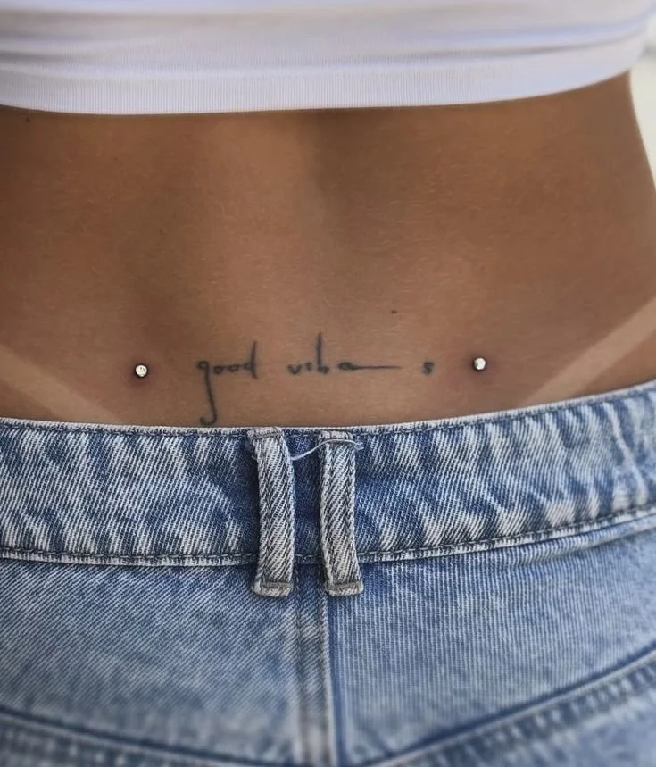Particolare di una persona con tatuaggio sul basso addome, che dice 'good vibes', indossando jeans e un top bianco.