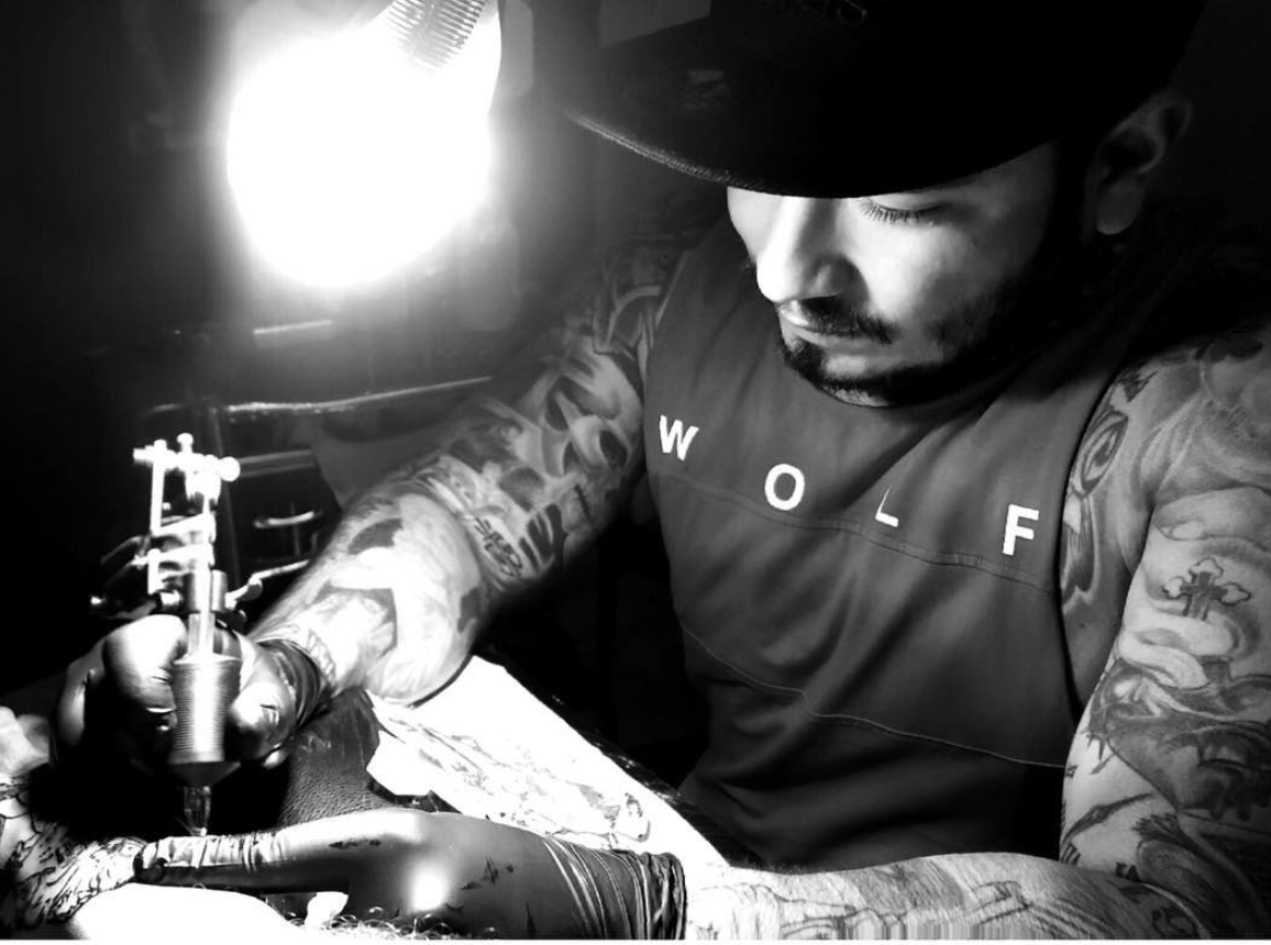 Uomo tatuato con cappello nero, TATUATORE, lavora in uno studio , illuminato da una lampada forte.