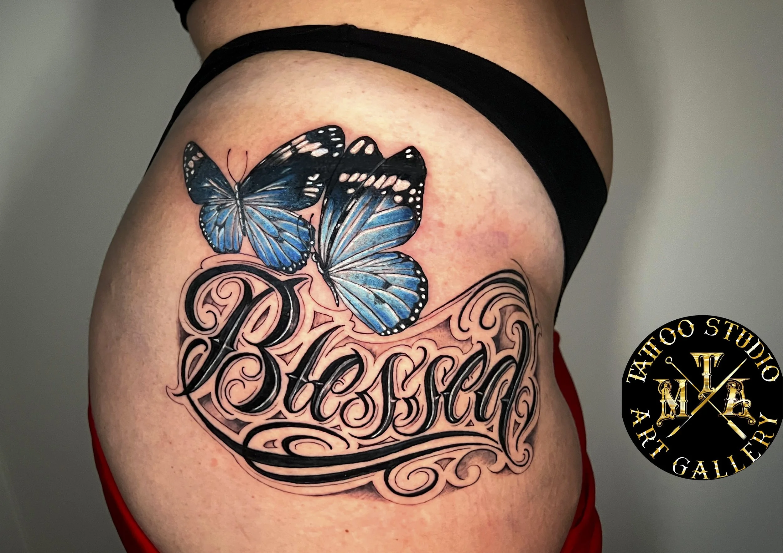 Tatuaggio con due farfalle blu e un testo decorative 'Blessed' sulla coscia di una persona.