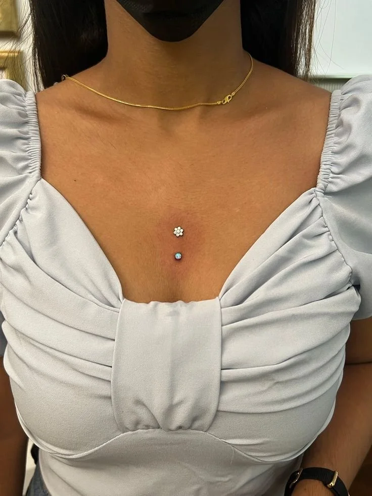 Close-up di una donna che indossa una maglia bianca con dettaglio arricciato, una collana sottile d'oro e due piercing al petto, uno a fiore e uno con una pietra blu.