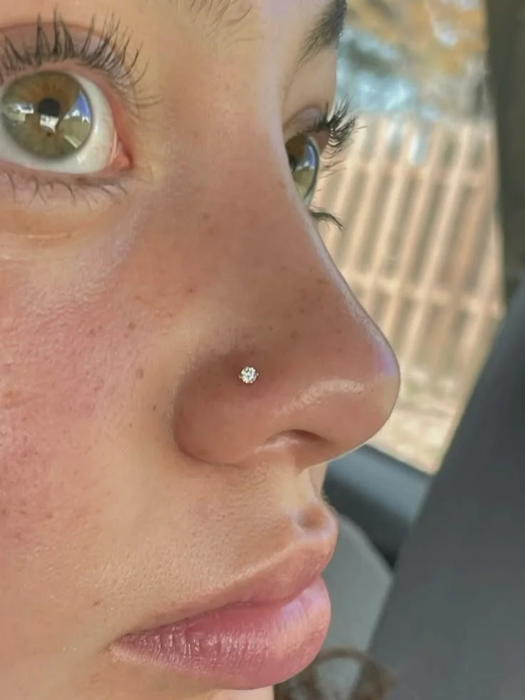 Primo piano del volto di una donna con piercing al naso, occhi castani, ciglia lunghe e pelle leggermente abbronzata.