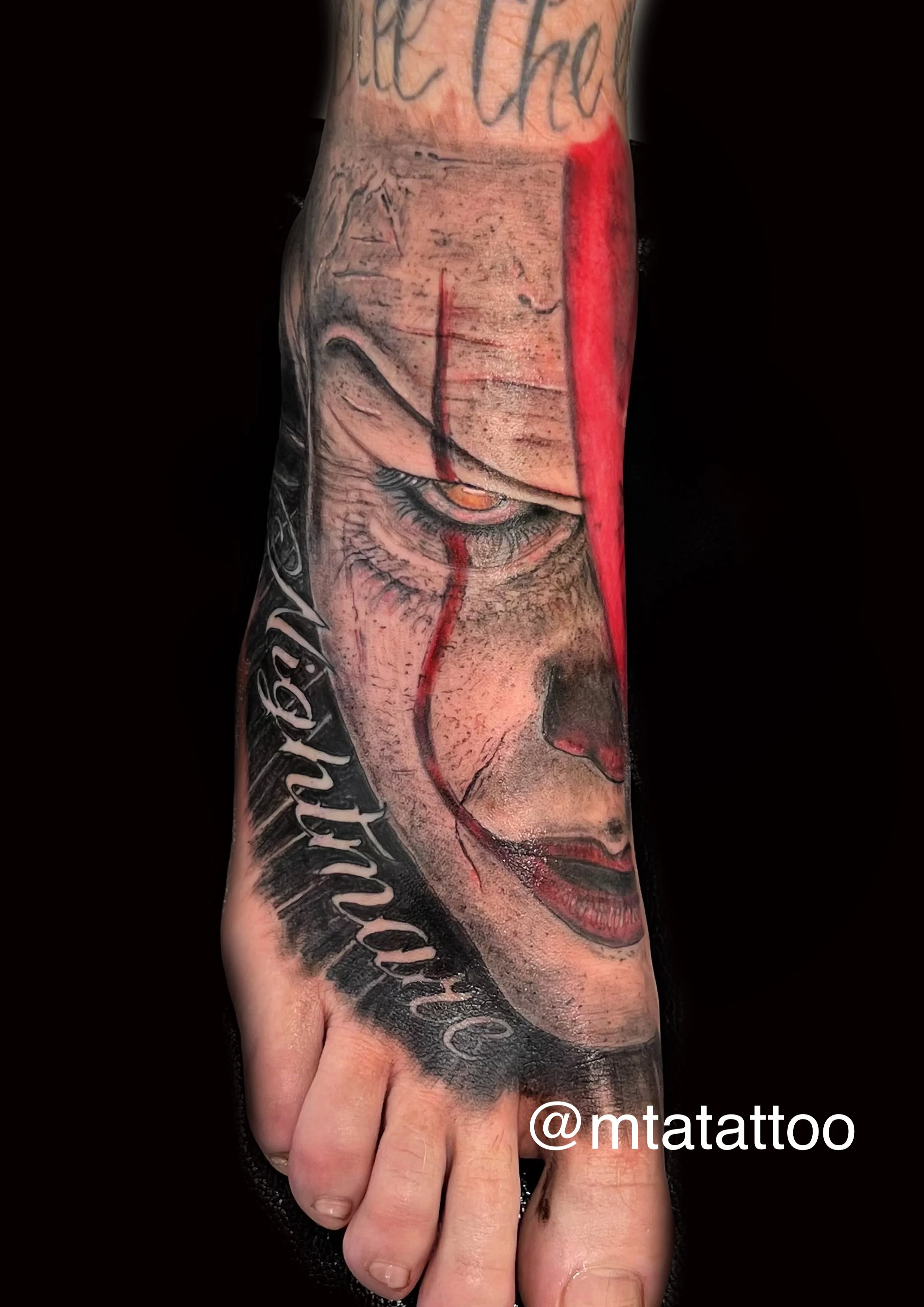 Tatuaggio realistico di un volto con dettagli di trucco e lacrime di sangue sul piede. La parte superiore del tatuaggio contiene testo scritto in corsivo e un tag di Instagram '@mtattoo'.