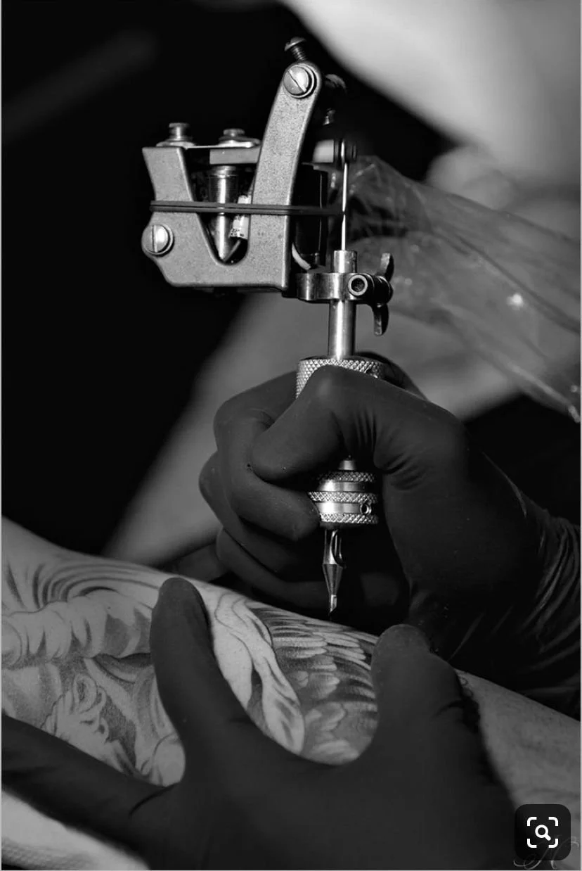 Una persona con guanti neri sta facendo un tatuaggio, usando una macchina per tatuaggi e un ago, su una parte del corpo che sembra essere un braccio con un disegno complesso.