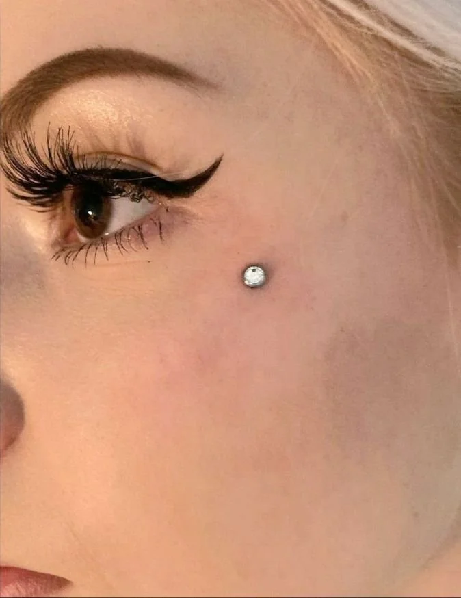 Primo piano di una donna con trucco occhi elaborato e un piercing con cristallo sotto l'occhio.