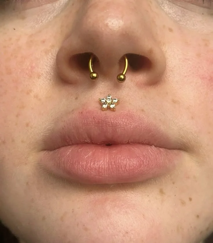 Primo piano di una persona con piercing al naso, con due anelli dorati e un piercing al septum con una decorazione a forma di farfalla.