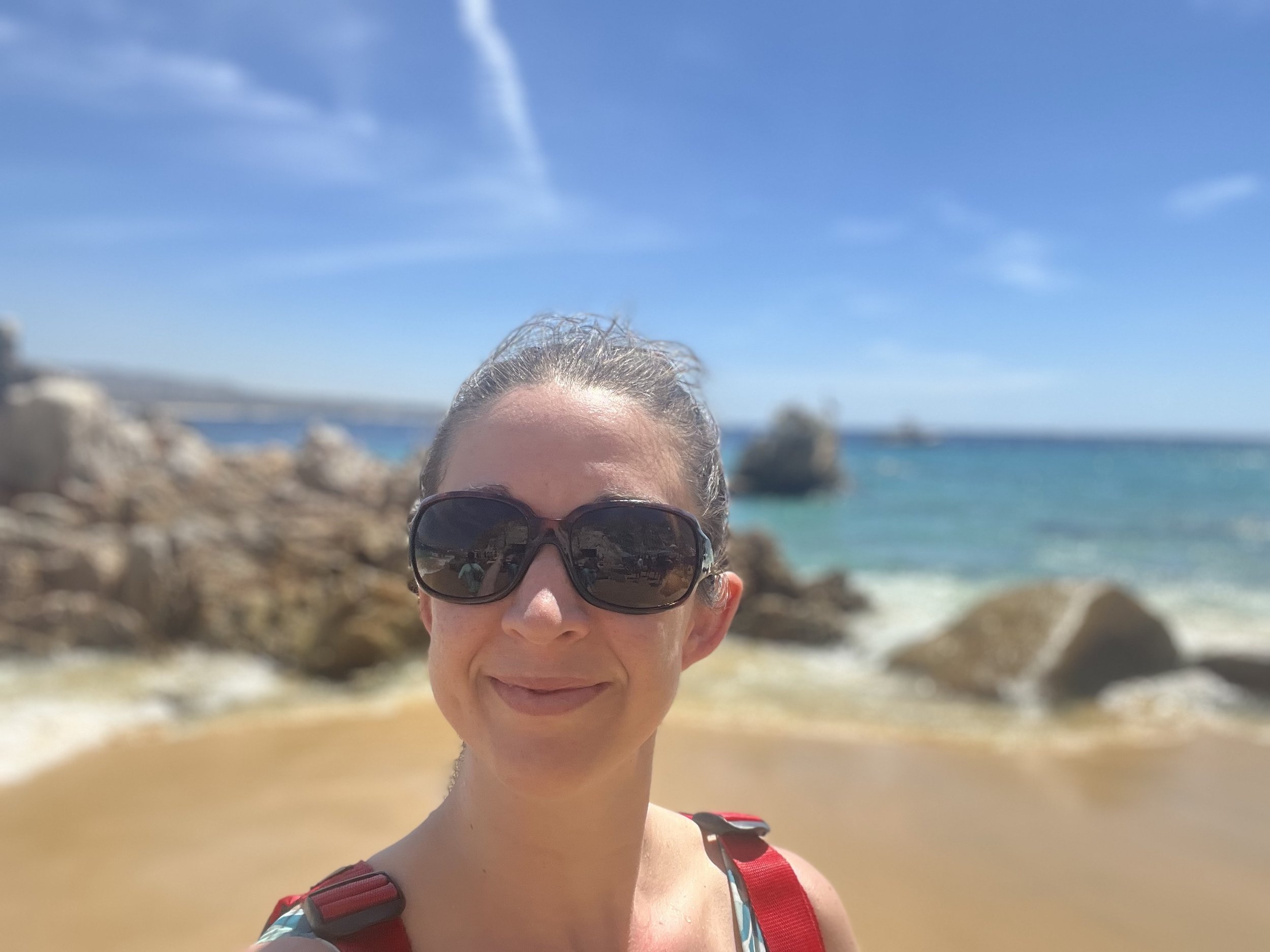 Woman with sunglasses smiling at the beach, rocks and ocean in the background under a blue sky.