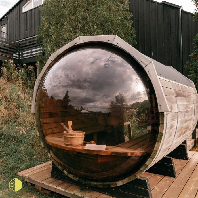A barrel-shaped outdoor sauna with a large glass front, reflecting the cloudy sky, situated on a wooden deck in a natural setting.