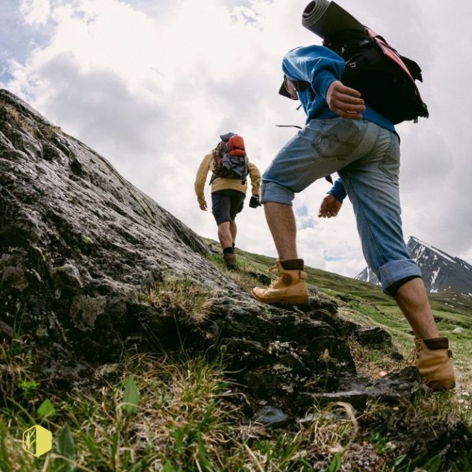 Two hikers climbing a grassy mountain trail with backpacks and hiking boots, with cloudy sky and snow-capped peaks in the background.
