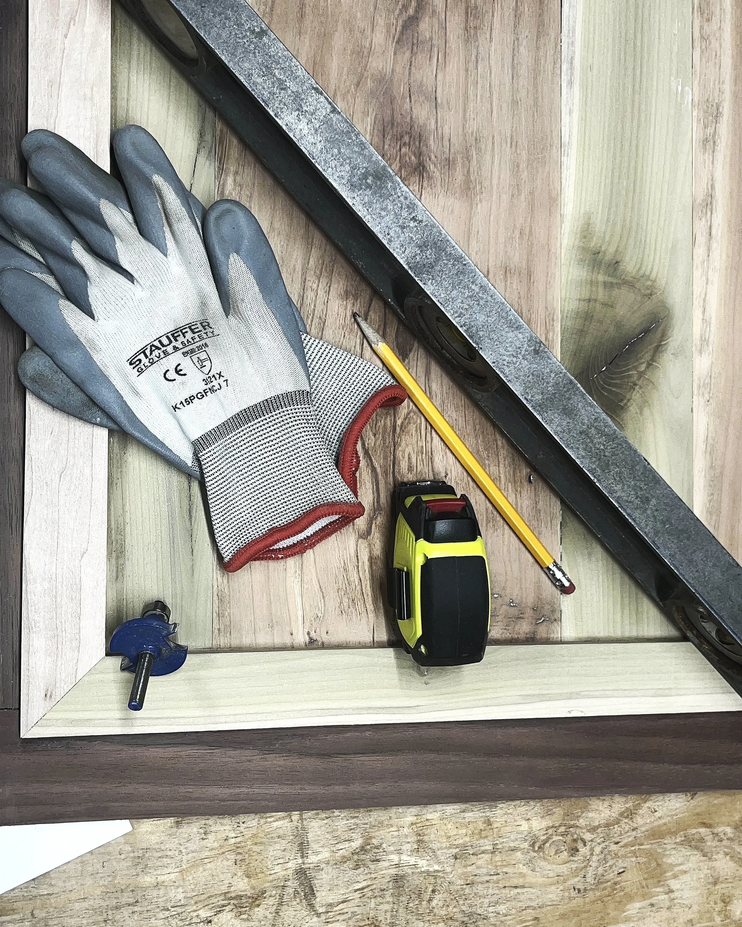 Flooring installation tools and materials, including a pair of work gloves, a level, a measuring tape, a yellow pencil, and a corner of wood framing, arranged on a wooden surface.